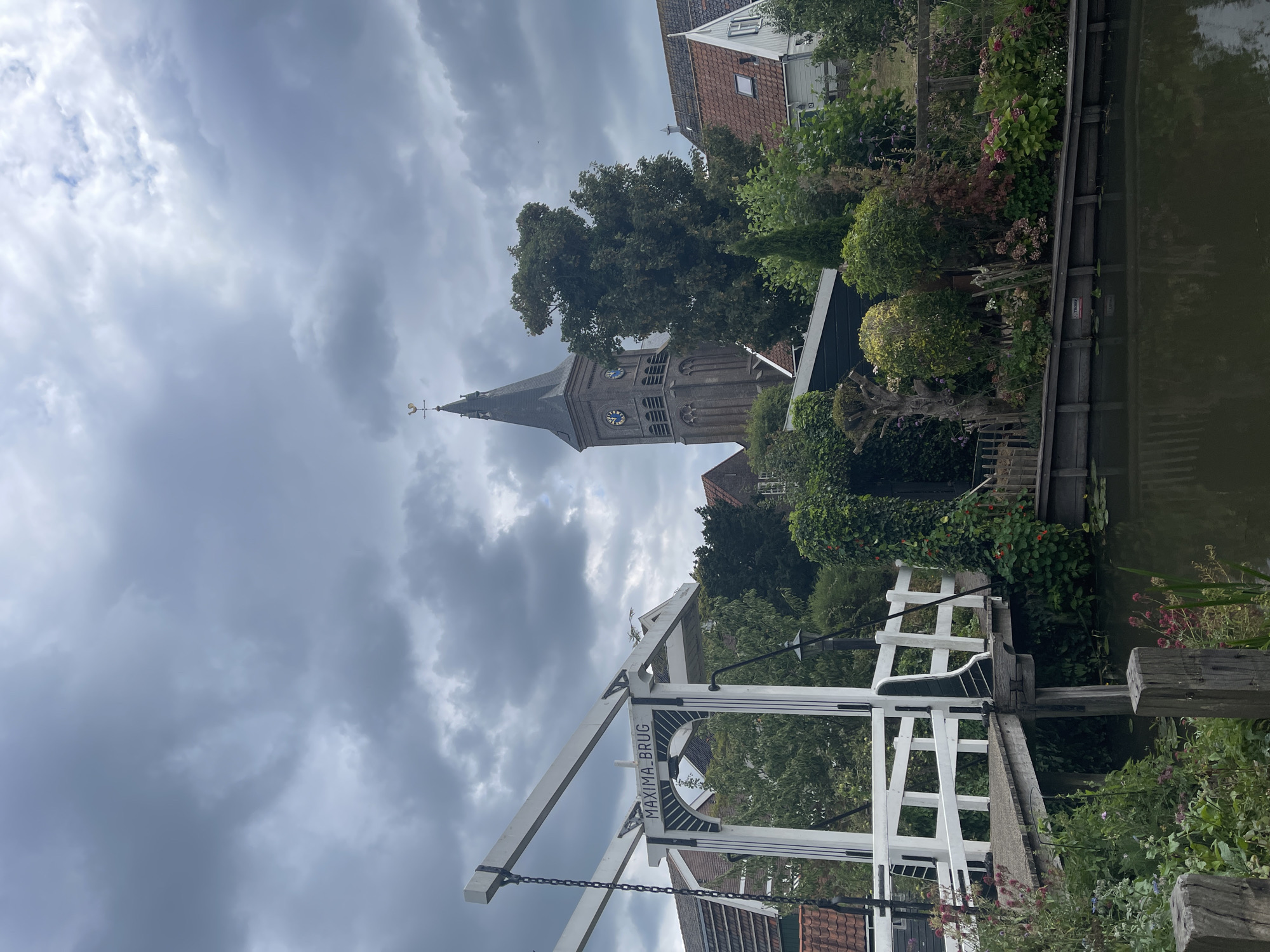 The image shows a picturesque canal scene in Marken, Netherlands. In the foreground, a white, arched drawbridge, labeled "MAXIMA BRUG," spans the dark water. The bridge is made of wood and has white railings. On the left side of the canal, lush green vegetation and flowers grow along the bank, with some red flowers visible.
In the middle ground, several houses with red-tiled roofs are nestled among trees. A tall, dark church tower with a clock face and a weather vane on top dominates the background, piercing a cloudy sky. The church tower appears to be made of brick. The overall atmosphere is serene and tranquil, characteristic of a Dutch village. The sky is overcast, with dramatic grey clouds suggesting a possibility of rain. The water in the canal reflects the sky and the surrounding greenery. There are no people or animals visible in the image.