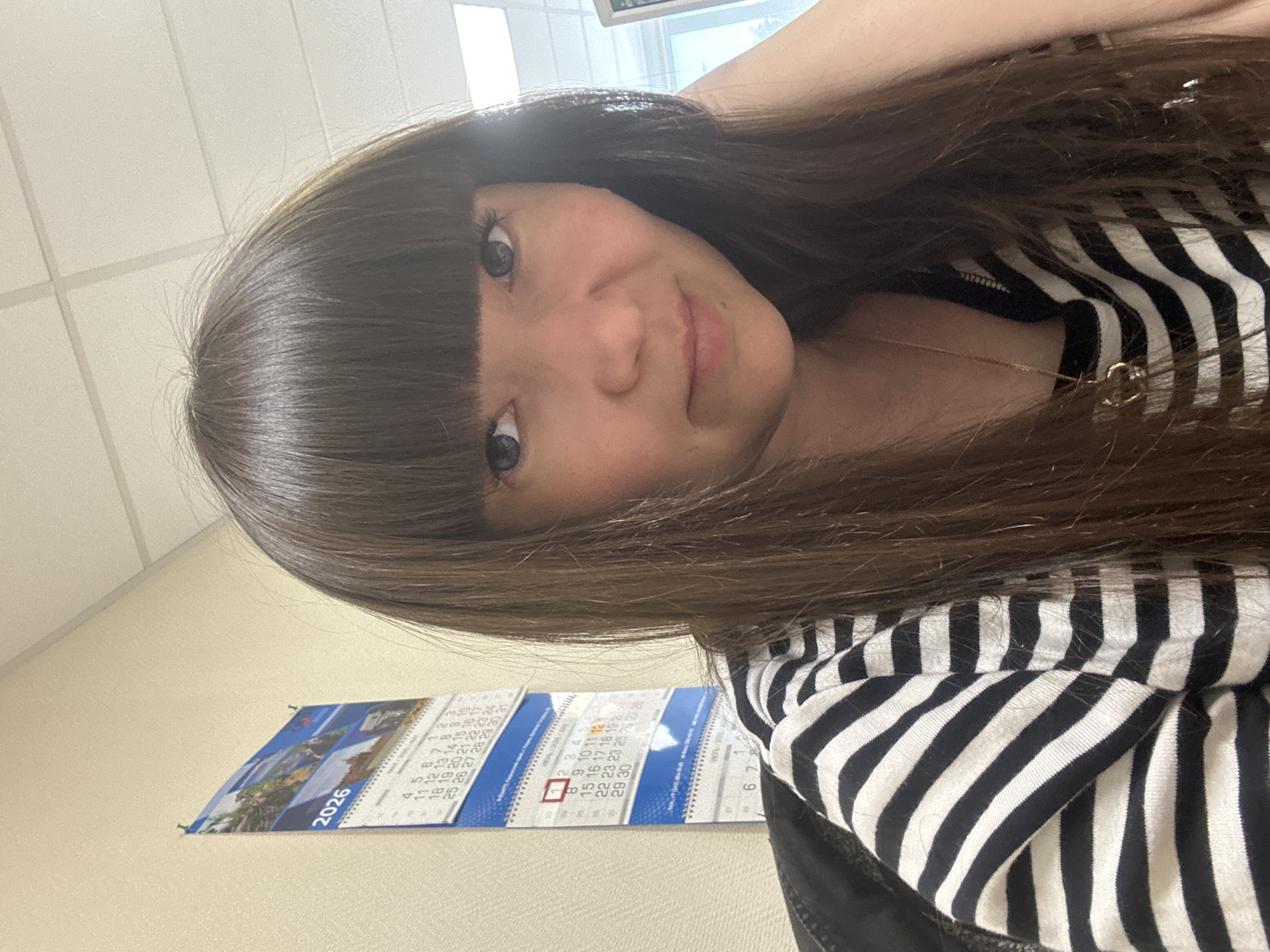 The image shows a woman with dark hair and bangs, wearing a black and white striped shirt. She is indoors, with a calendar hanging on the wall behind her. The calendar shows the year 2026. The woman is looking at the camera. Based on the partial OCR on the calendar, it includes telephone prefixes +7 (34241) which corresponds to the city of Chaikovsky, Russia.