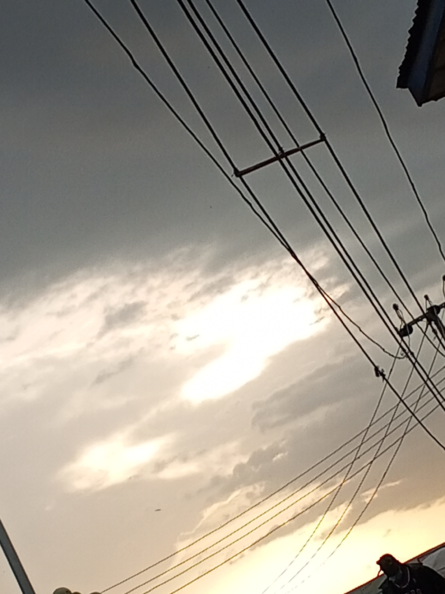 The image shows a cloudy sky with the sun shining through the clouds, creating a bright spot. Several power lines are visible, crisscrossing the sky. A portion of a building is visible in the top right corner. In the lower right corner, there is a person wearing a dark jacket and a cap. The location is likely in Gwong, Nigeria, based on the prompt. The scene depicts an ordinary urban landscape with utilities and a cloudy sky at dusk or dawn.