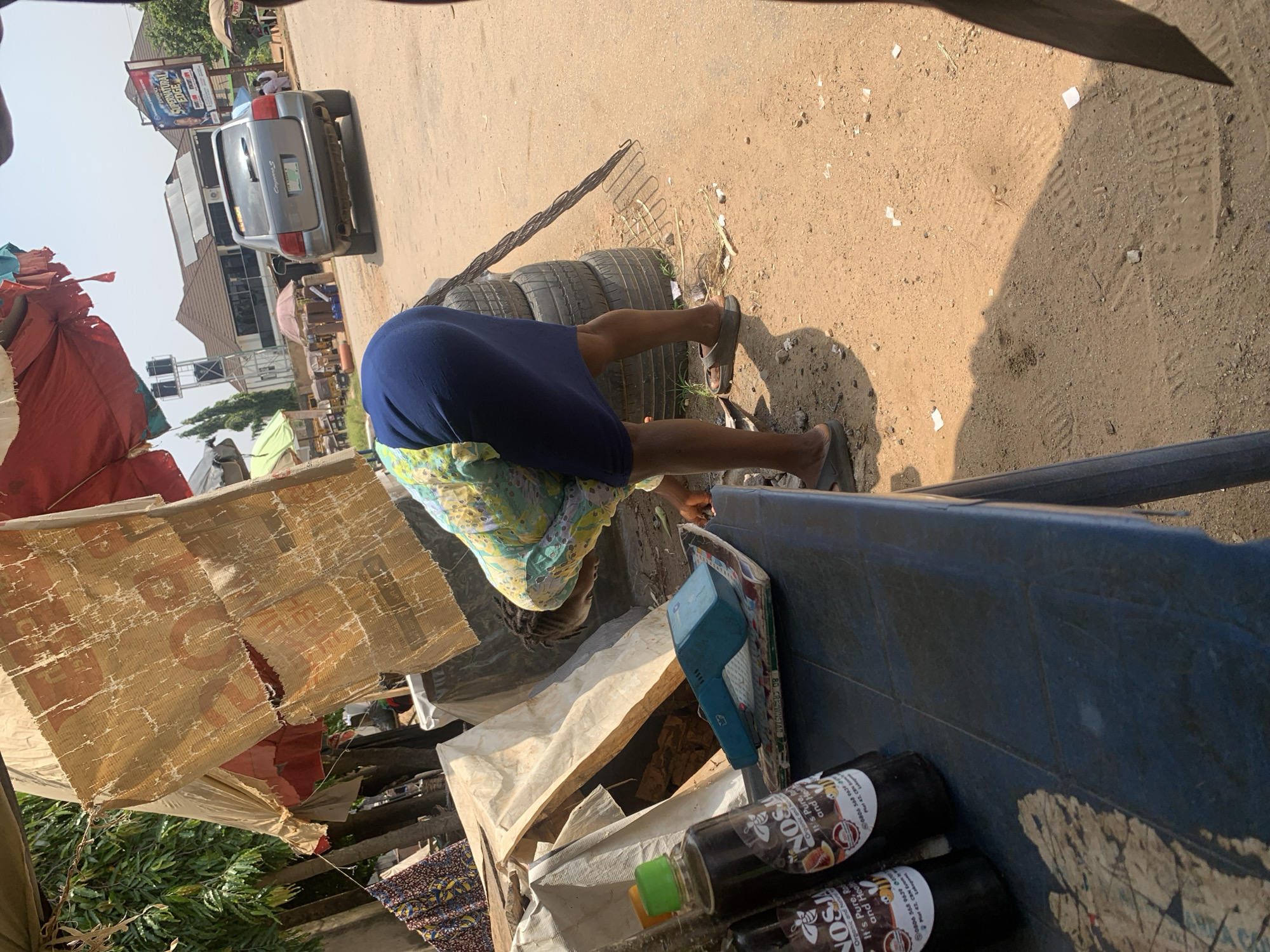 In a sun-drenched street scene in Abuja, Nigeria, a woman is bent over, her attention focused on items on a blue plastic table. She wears a blue skirt and a patterned yellow and green top. On the table, two dark bottles with labels are visible, along with a teal-colored electronic device. Behind her, a stack of tires and some discarded materials are present. In the background, a grey SUV is parked on a dusty road, and various buildings and market stalls are scattered throughout the area, suggesting a busy commercial or residential district. The overall environment is casual and somewhat cluttered, typical of a street market or informal trading post.