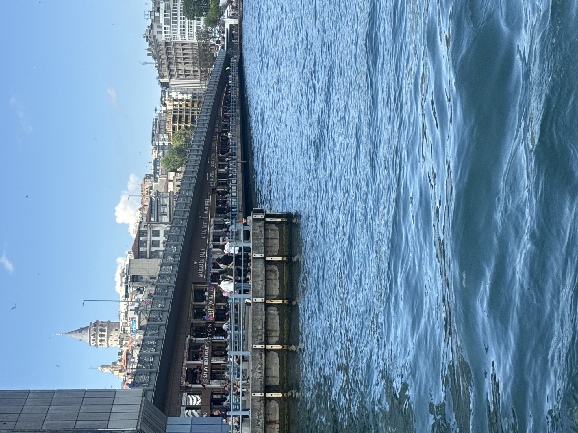 The image shows the Galata Tower and the Galata Bridge in Istanbul, Turkey. The view is looking toward the tower across the Golden Horn. The Galata Bridge is lined with restaurants and people are visible dining in these establishments. The water of the Golden Horn is in the foreground. The sky is blue. Buildings can be seen along the waterfront.