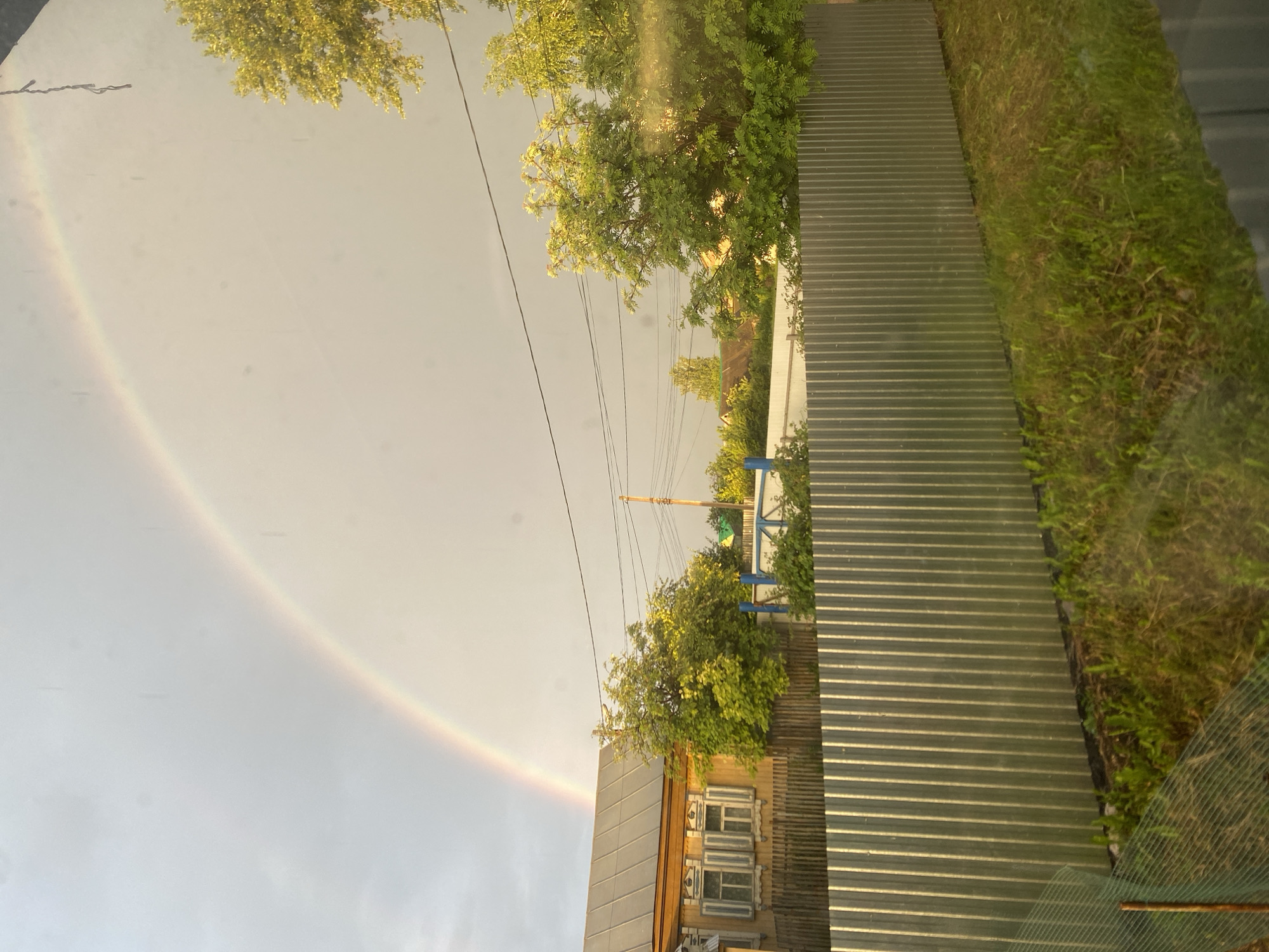 In the city of Chirsha-Tartysh, Russia, a rainbow arches across a sky that is transitioning from overcast to clearer. In the foreground, a corrugated metal fence runs horizontally across the frame, with a patch of green grass and some dry vegetation in front of it. Behind the fence, a rustic wooden house with white-shuttered windows is visible. Several trees with vibrant green foliage are scattered around the property. Power lines are strung across the sky, supported by a utility pole. The overall scene depicts a rural or suburban environment under a sky marked by the presence of a rainbow after a rain shower.