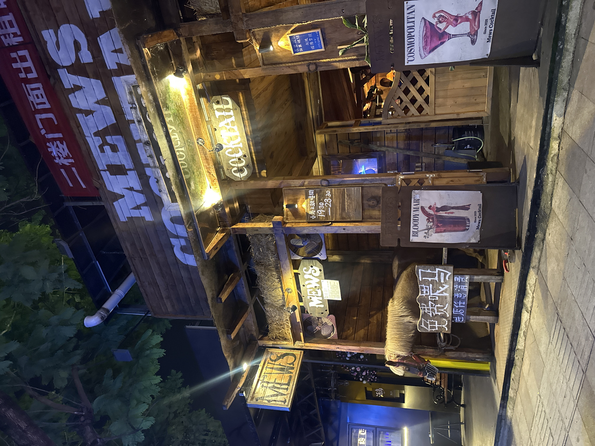 The image shows the exterior of a bar or restaurant named "MEWS" in the Chongqing High-tech Zone, China. The establishment has a rustic, wooden facade with prominent "MEWS" signage. Above the entrance, a red banner with Chinese characters is visible. To the left of the entrance, a wooden sign reads "MEWS" and another sign below it has Chinese characters and the text "COCKTAIL". A live horse is present inside a partially enclosed area to the left of the entrance, with a sign in front of it that reads "免费骑马" (Free Horse Riding). To the right of the entrance, there are posters advertising "BLOODY MARY" and "COSMOPOLITAN" cocktails, both also referencing "Mews Cocktail" and a "Since 2025" date. The scene is lit by artificial lights and appears to be at night. The ground in front of the establishment is paved with cobblestones.