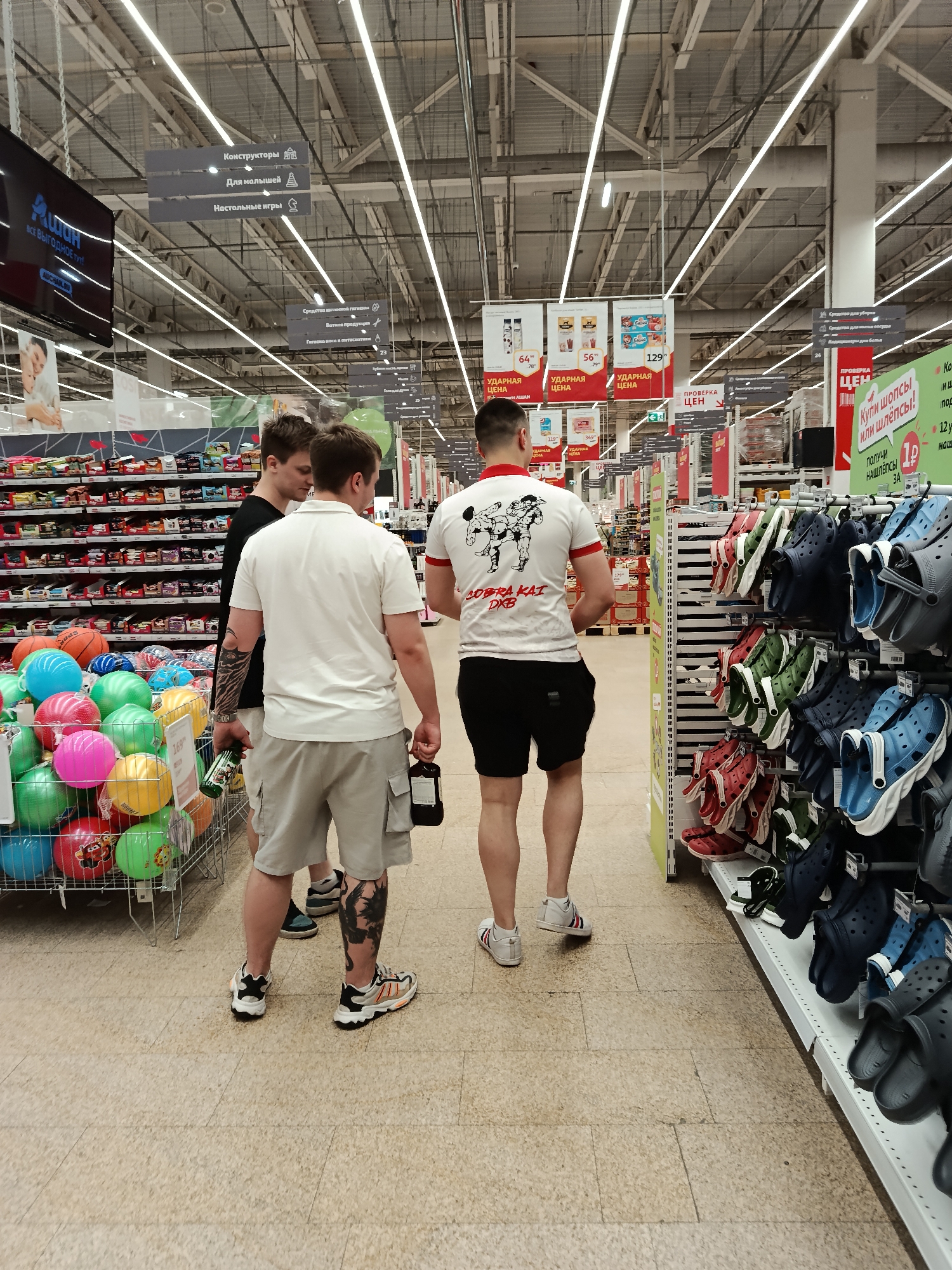 The image shows three young men in the Auchan supermarket in Moscow, Russia. The man in the center wears a white polo shirt and gray cargo shorts, while the man to his left wears a black t-shirt and shorts. The man to the right wears a white t-shirt with a "Cobra Kai DXB" design on the back and black shorts. They are standing in an aisle with shelves of colorful plastic balls on the left and shoes on the right. The man in the center carries a dark bottle in his right hand and a green bottle in his left hand. Overhead signs are visible, indicating the locations of different sections in the store, such as "Constructors," "For Kids," and "Board Games." Price tags and promotional materials are also visible on the shelves.