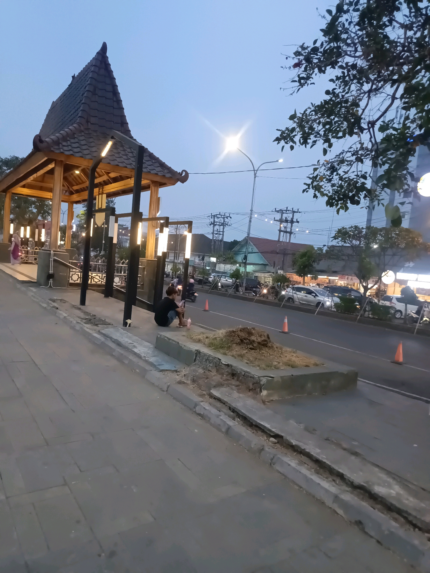 The image depicts a street scene in Lubuklinggau, Indonesia, during dusk. A young person is seated on the edge of a sidewalk next to a planter box, appearing to be looking at their phone. The sidewalk is paved with stone tiles. Adjacent to the sidewalk is a road with several vehicles, including cars and motorcycles, in motion. On the other side of the road, buildings with illuminated storefronts are visible, suggesting commercial activity. A distinctive traditional Indonesian-style gazebo with a tiled roof stands on the left side of the frame, illuminated by warm lights. The sky is a pale blue, indicating the transition from daylight to night. The overall atmosphere is that of a moderately busy urban environment in the early evening.