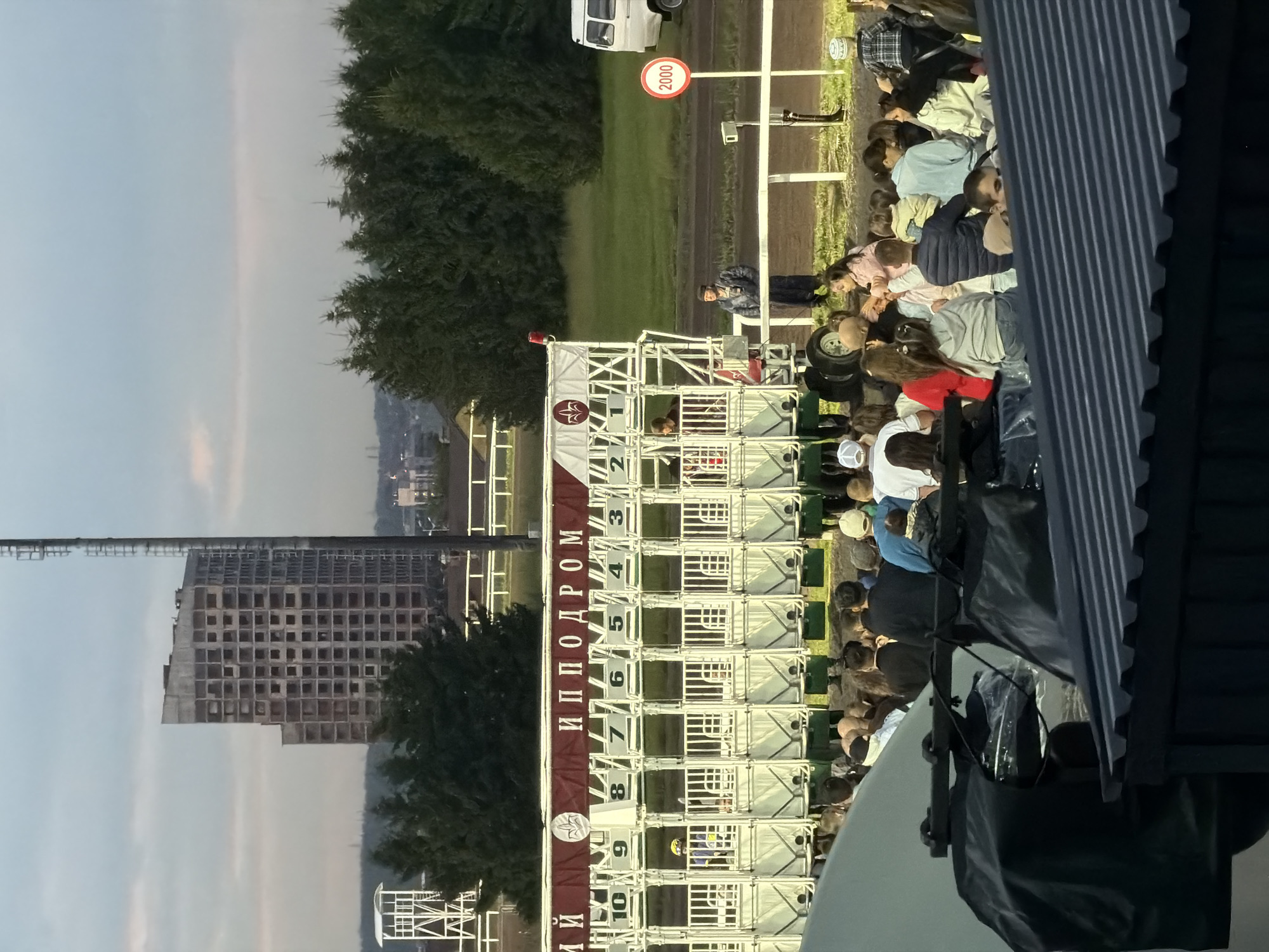The image depicts a horse racing event at a hippodrome. In the foreground, a crowd of spectators is visible, some seated and others standing, watching the event. To the left, a dark, covered structure with a corrugated roof is present. Behind the spectators, a starting gate with numbered stalls is set up on a track. Several horses with jockeys in colorful attire are positioned in the stalls. A banner above the gate reads "ИППОДРОМ" in Cyrillic. In the middle ground, a person in a dark coat stands near a white railing. A white van is parked on the right side of the image. A red circular sign with the number "2000" is visible on a pole. The background features a line of trees and a tall, unfinished building with a grid-like facade, suggesting an urban or semi-urban setting. The sky is overcast with diffused light.