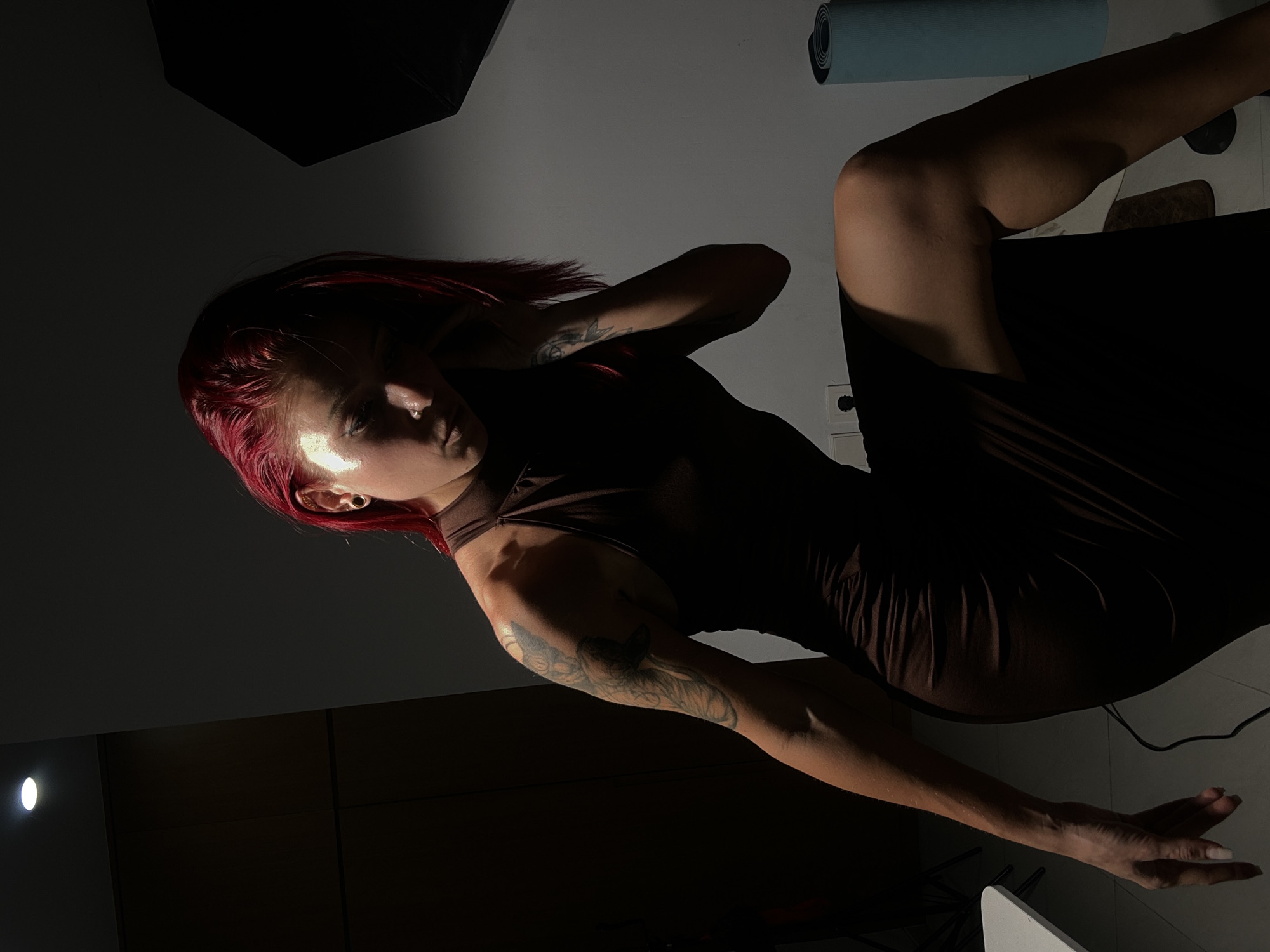 A woman with red hair and tattoos on her arms is sitting on a dark surface in a dimly lit room. She is wearing a brown dress. A rolled-up blue object is visible in the background. The location is not evident in the image.