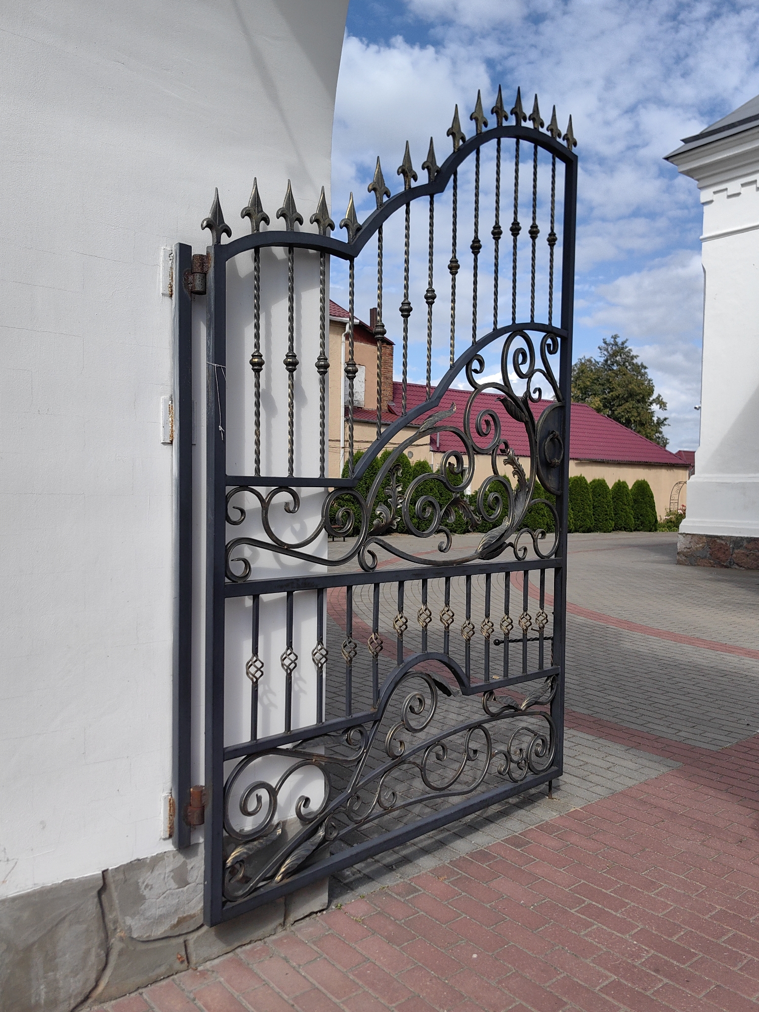 This is an outdoor, eye-level, medium shot photograph of an open, ornate, black metal gate. The gate features decorative scrollwork, twisted metal bars, and pointed finials at the top. It is attached to a white, textured wall on the left by a rusty hinge. The gate is set into a pathway paved with red and gray interlocking bricks. In the background, a building with a red roof is visible, partially obscured by dark green evergreen bushes. The sky above is blue with scattered white clouds. The location is identified as the St. Nicholas Church in Pastavy, Belarus. There are no people visible in the image.