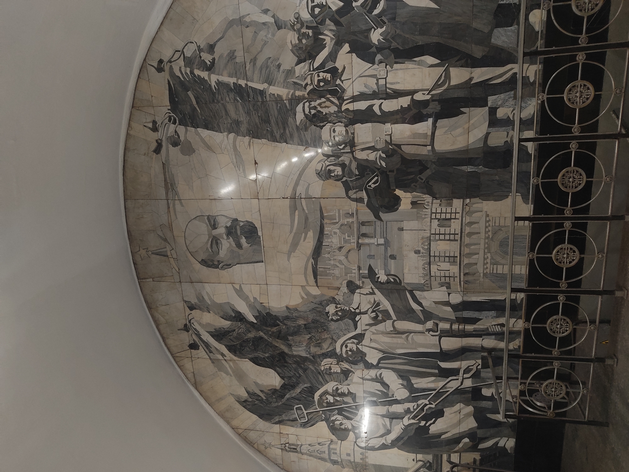 A low-angle shot depicts a large, rounded mosaic mural set within a metro station of Unknown, Unknown. The mural, rendered in shades of gray and white, is composed of numerous tiles. The subject of the mural appears to be historical, featuring a portrait of Lenin, along with groups of people, banners, and buildings. A metal railing with decorative circular elements is situated to the right of the mural. The curved edge of the wall on which the mosaic is mounted is visible on the left side of the frame.