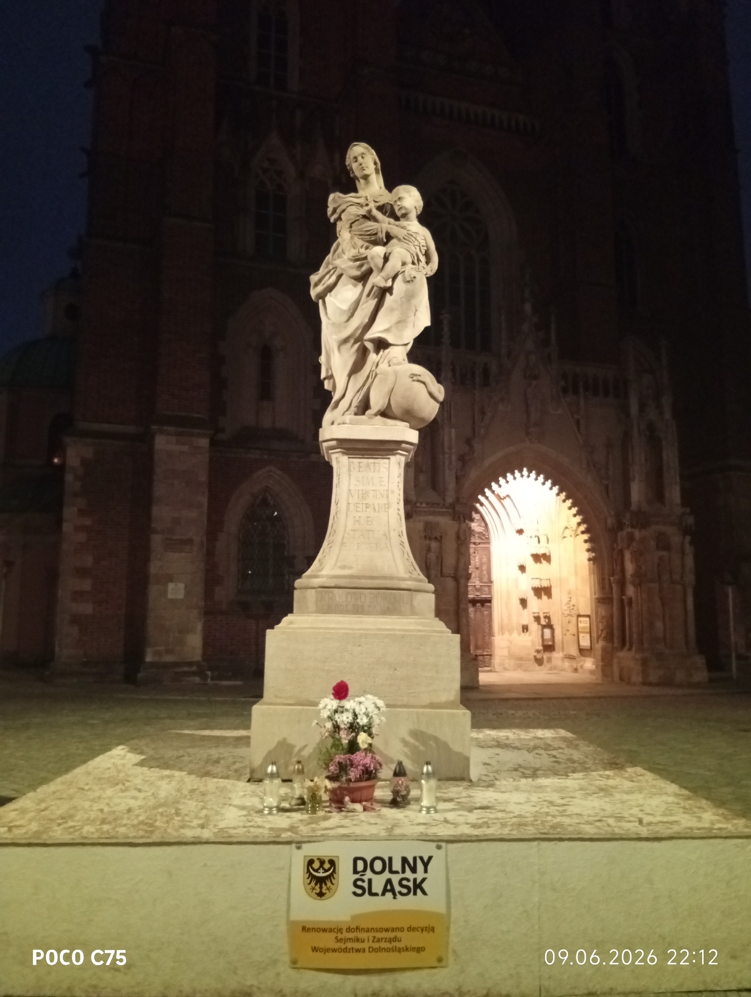 The image captures a nighttime view of a statue of the Virgin Mary and Child in front of a Gothic-style building, likely a cathedral, in Wrocław, Poland. The statue, carved from light-colored stone, stands on a pedestal with inscriptions. At the base of the pedestal, a small bouquet of flowers and two votive candles are placed. Below the flowers, a yellow sign with the text "DOLNY ŚLĄSK" and a coat of arms is visible, along with smaller text indicating a renovation financed by the Lower Silesian Voivodeship Board. The building behind the statue features ornate architectural details and a brightly lit arched entrance. The sky is dark blue, suggesting it is night or late evening. The image also includes a watermark in the lower left corner reading "POCO C75" and a timestamp in the lower right corner reading "09.06.2026 22:12".