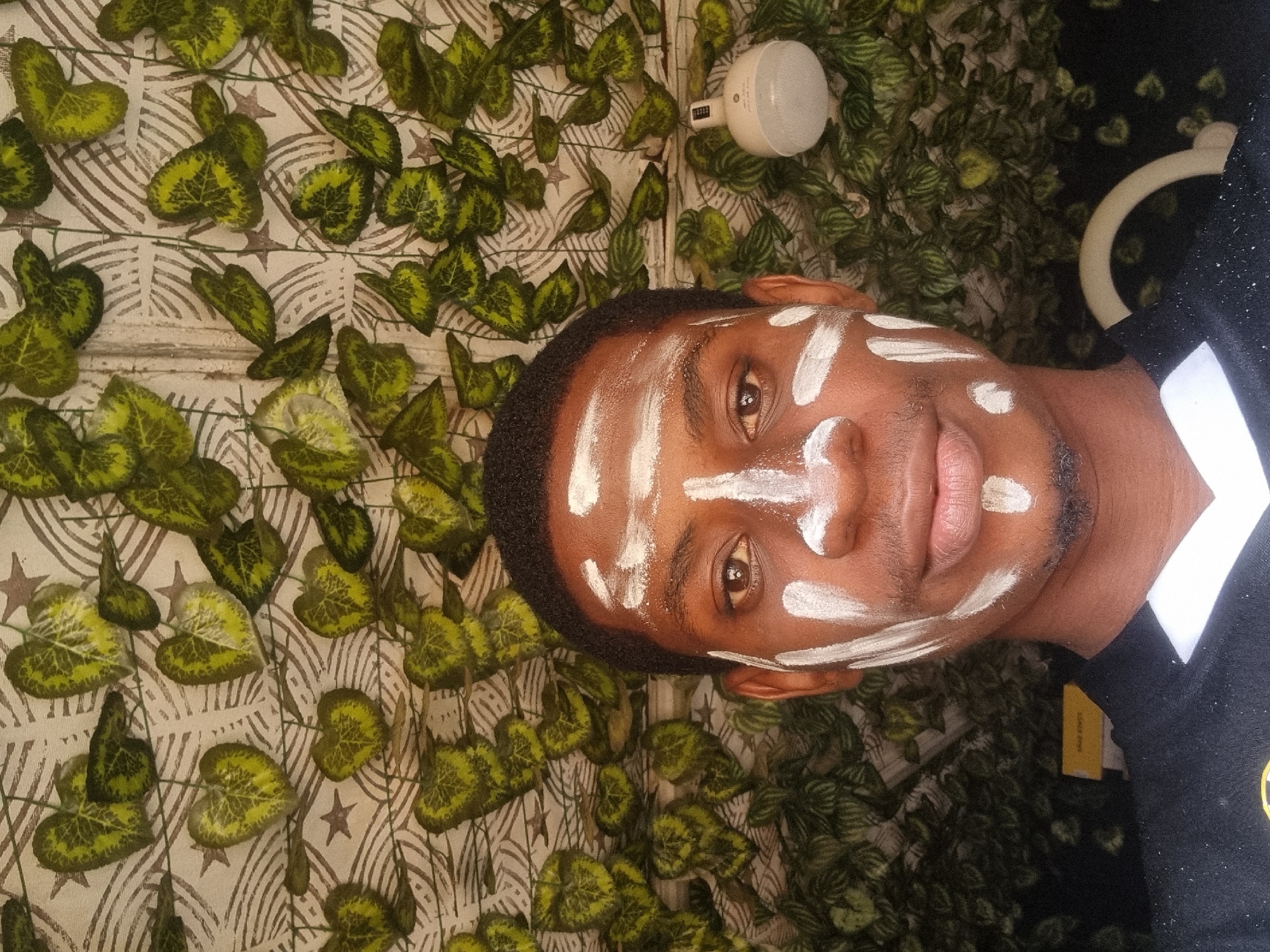 A headshot of a Black male with white streaks on his forehead, nose, and cheeks. The man is wearing a dark collared shirt with a white collar and some kind of logo on the right breast. The background consists of a beige patterned wall with green leaf designs, and a light bulb is visible near the top of the frame. The location cannot be determined from the information given.