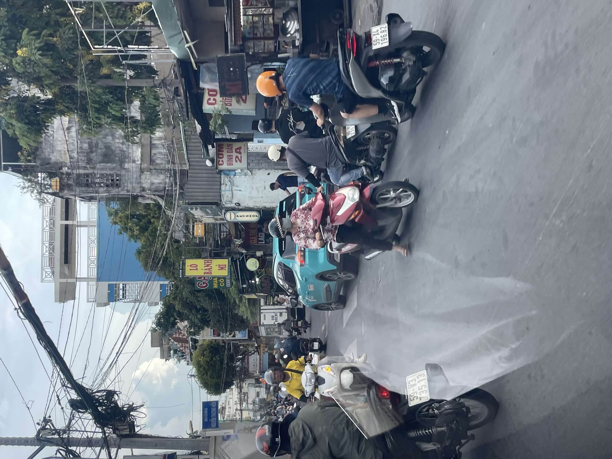 The image depicts a bustling street scene in Thủ Đức City, Vietnam, likely during the daytime under an overcast sky. The foreground features the rear of a motorcycle with a license plate reading "59-V3 735.13." The road is filled with various vehicles, predominantly motorcycles, navigating through traffic. Several individuals are visible riding motorcycles, some wearing helmets and face masks, common attire in this region.
In the middle ground, a bright teal car is prominent, indicating a mix of vehicle types. A woman on a red motorcycle is captured in motion. Behind them, a man in a yellow shirt on a white scooter is a focal point, appearing to be actively participating in the traffic flow. Further back, more motorcycles and a truck are present, contributing to the sense of a busy urban environment.
The background reveals the typical architecture of Vietnamese cities, with multi-story buildings lining the street. Some structures appear aged and weathered, while others show signs of modern construction, including a building with a blue facade and balconies. Overhead, a dense network of electrical wires crisscrosses the sky, a characteristic feature of many Asian urban landscapes. Street signs are visible, some in Vietnamese script, suggesting commercial establishments and directional information. The overall atmosphere is one of active daily life and movement within a dense urban setting.