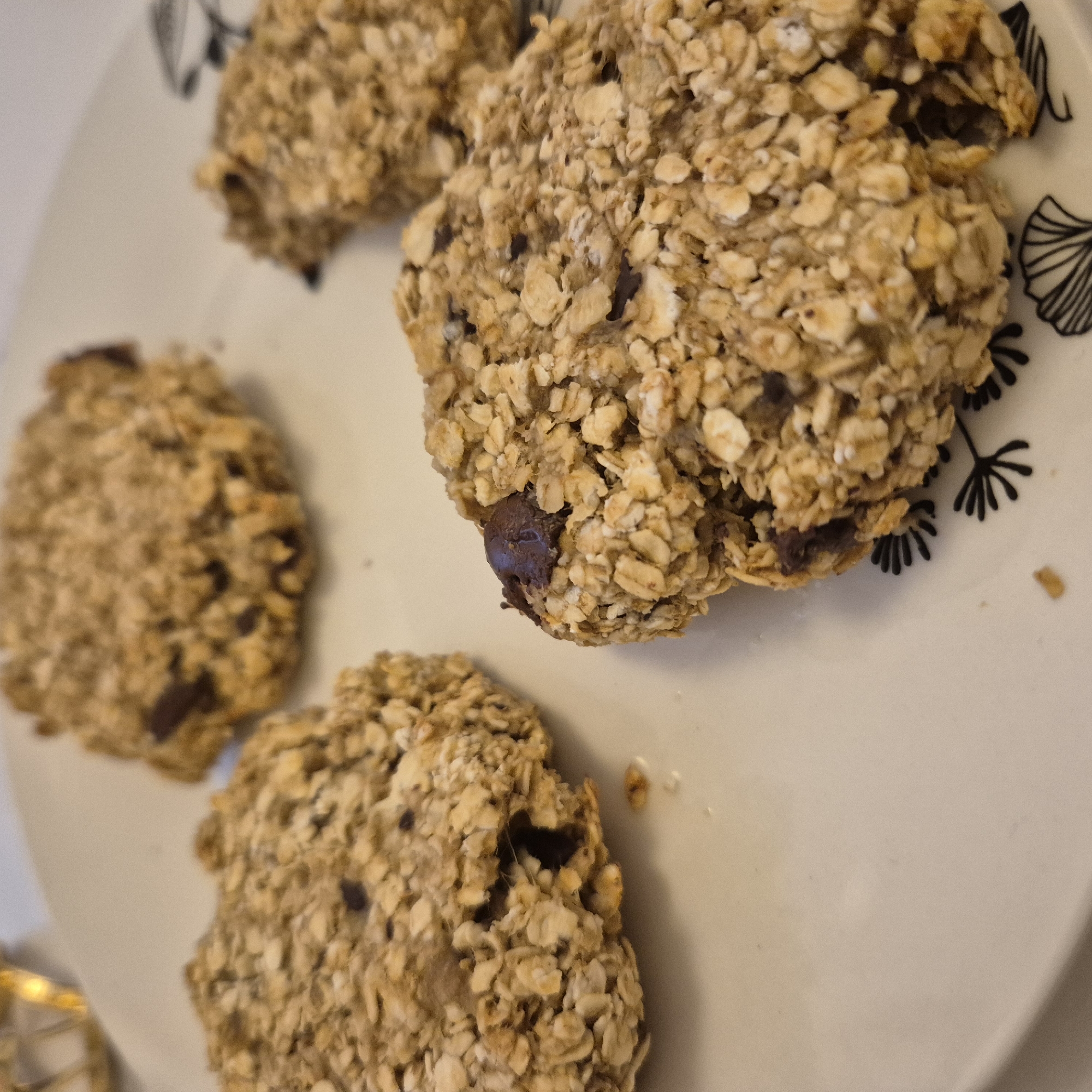 The image shows four oatmeal chocolate chip cookies on a white plate with a black floral pattern on the upper right corner. The cookies are irregularly shaped and appear to be homemade. Some of the chocolate chips are visible. A portion of a gold-colored object is visible in the lower-left corner. The location is not discernible from the image.