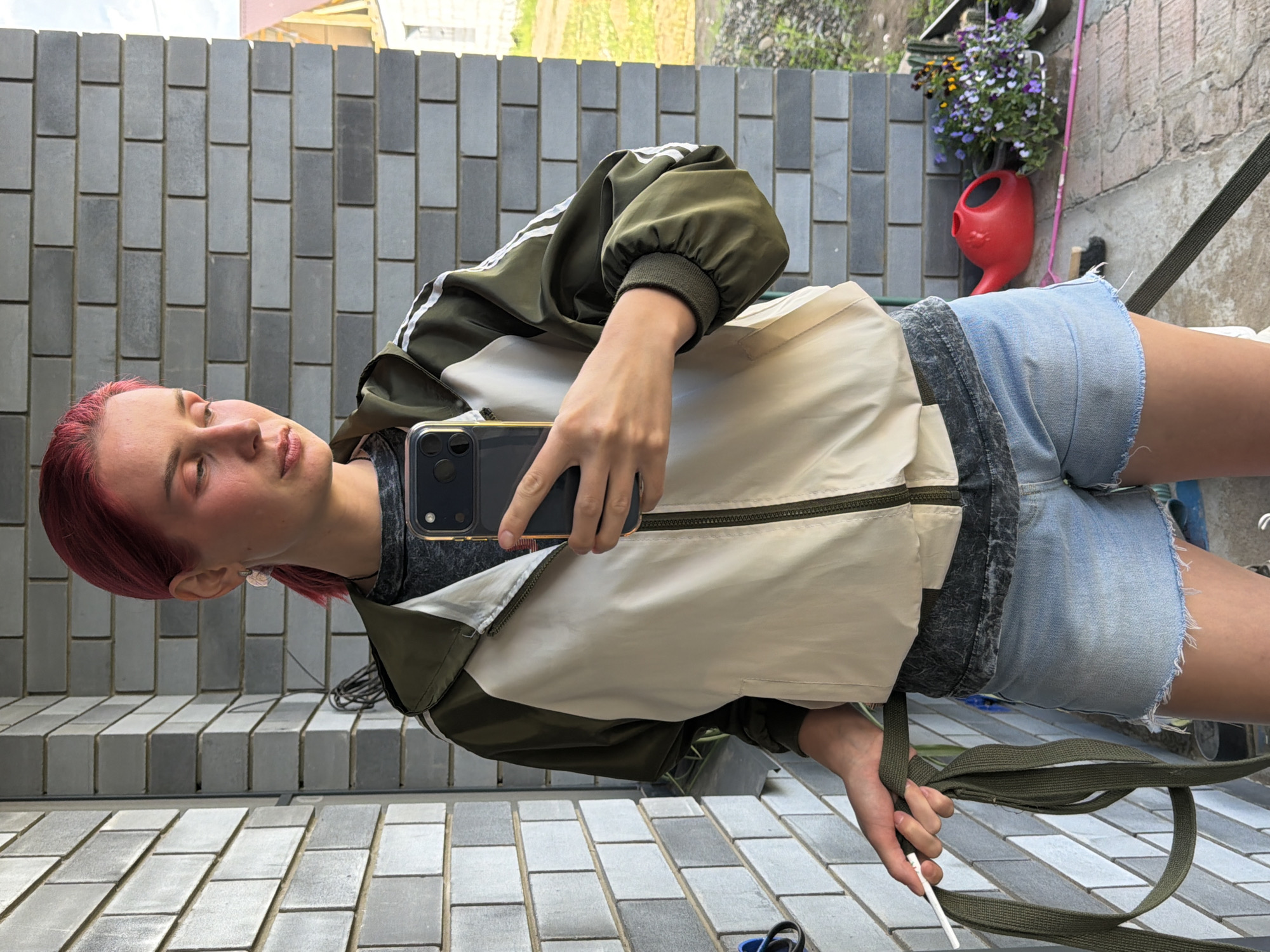 This is a vertical, medium shot photograph taken outdoors in Chaikovsky, Russia. The subject is a young person with short, vibrant red hair, who is looking into a smartphone held in their right hand, presumably taking a selfie in a mirrored surface, as indicated by the reflection of the background. They are wearing a green and cream-colored windbreaker with white stripes on the sleeves and light-wash denim shorts. The background consists of a grey brick wall and a tiled surface. In the upper right portion of the frame, a red watering can with a distinctive face-like design is visible, hanging near a planter filled with purple and yellow flowers. The lighting suggests it is daytime, possibly sunny, given the bright reflections. The person's expression appears relaxed and neutral. There is no visible text in the image.