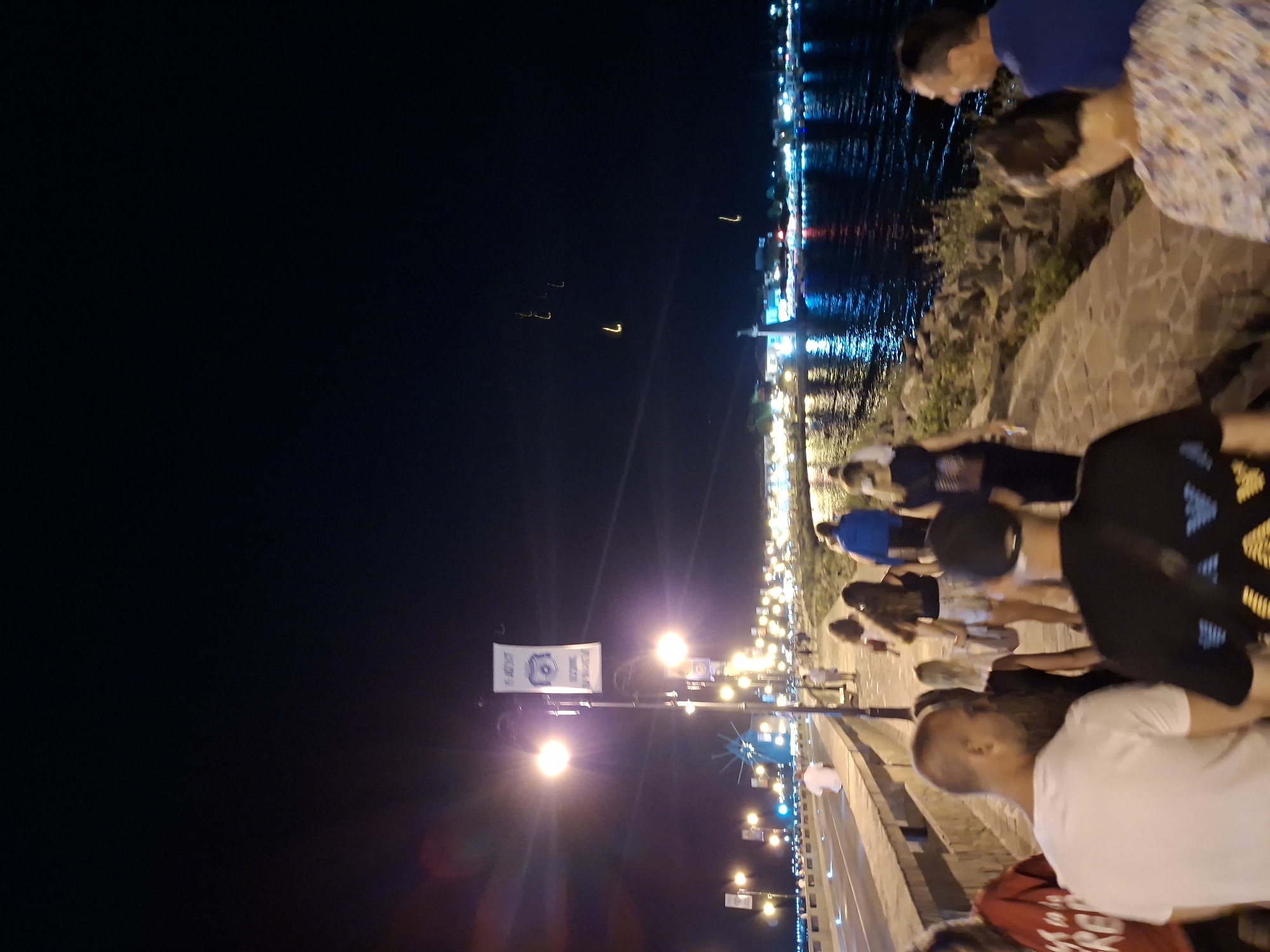 The image depicts a nighttime scene along a waterfront promenade. In the foreground, a group of people are walking away from the camera. The promenade is paved with stone and lined with a low wall. Beyond the wall is a body of water, reflecting the lights of a town or city in the distance. The town is illuminated with numerous lights, including streetlights and building lights, creating a vibrant glow on the water. Along the promenade, there are streetlights illuminating the path. Further in the distance, a lighthouse or tall structure is visible, and there are also some faint, glowing objects in the sky, possibly fireworks or aerial lights. The overall atmosphere is lively and suggests a public gathering or event at night.