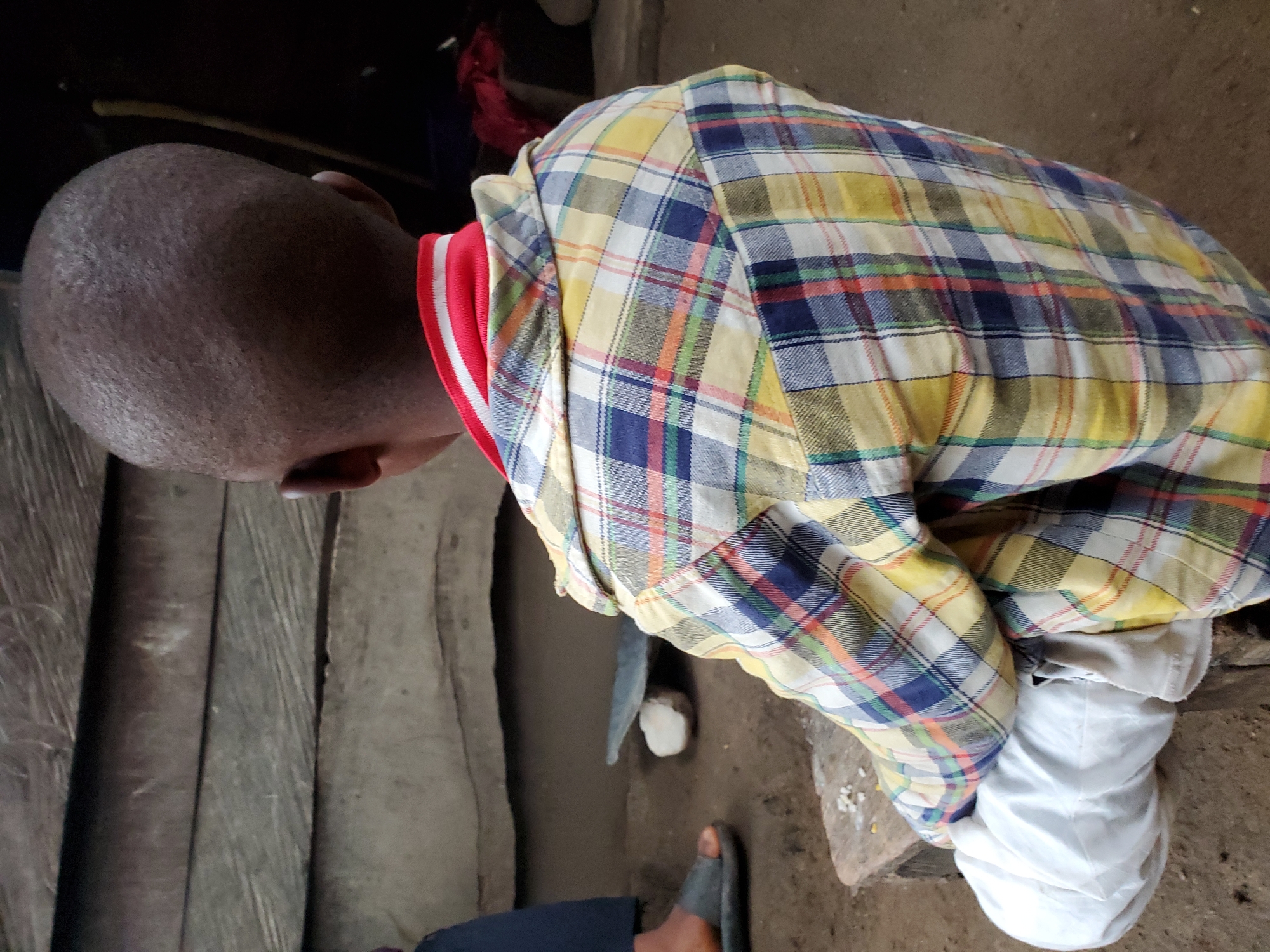 A man is sitting on a low wooden surface in Mararaba, Nigeria. He is wearing a plaid shirt with red trim at the neck and a white garment covering his legs. The man has a shaved head. The setting appears to be a simple interior with wooden walls. A sandal is visible on his foot.