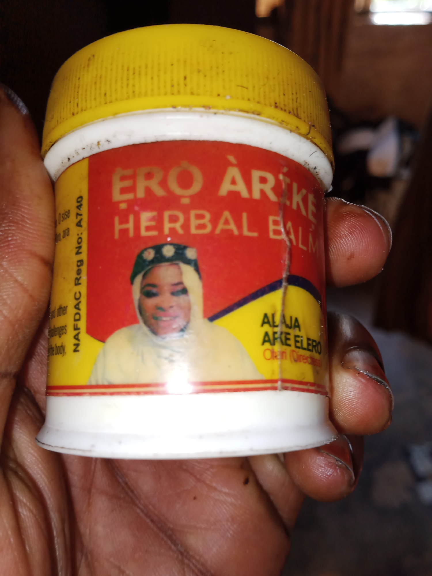 A close-up shot shows a person's hand holding a small, white plastic container with a yellow screw-on lid. The container has a red label with yellow text and a picture of a woman wearing a headscarf and smiling. The text on the label appears to be in a language other than English and includes "ERO AREKE HERBAL BALM" and "NAFDAC Reg No: A740". The background is blurred and appears to be an indoor environment with wooden structures, possibly in Jauro Yinu, Nigeria. The lighting is dim, suggesting it may be indoors during daylight or artificial light.