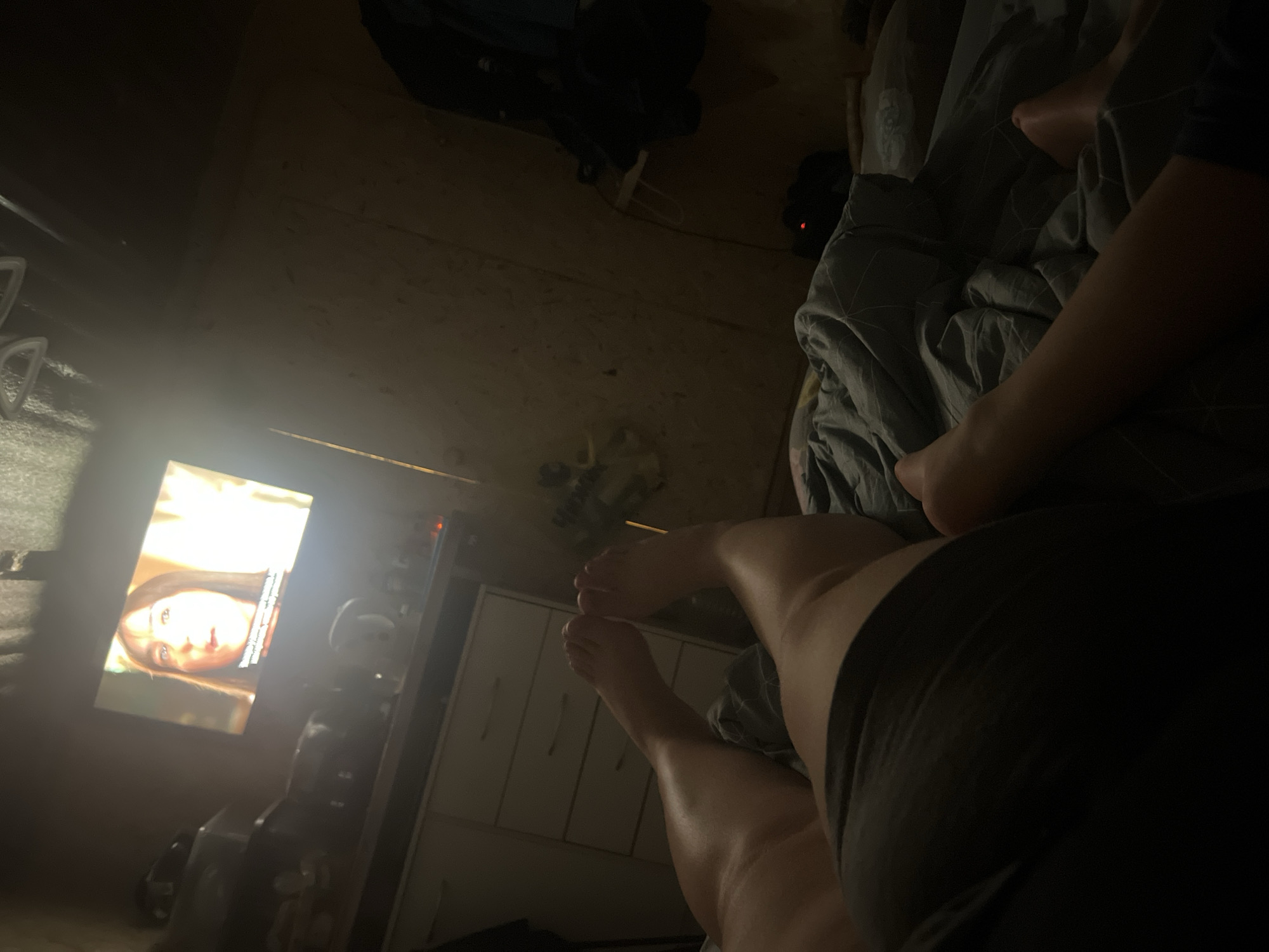The image depicts a person lying in bed watching television. The location is not discernible as Chaikovsky, Russia.
Subjects: A person is visible from the mid-thigh down to their feet.
Objects: A bed with grey patterned bedding, a television displaying a woman's face, a dresser with drawers, a fan, and various dark objects are visible.
Actions: The person is lying in bed watching television.
Interactions: The person is interacting with the television.
Environment: The room appears to be dimly lit and interior to a bedroom or living area.