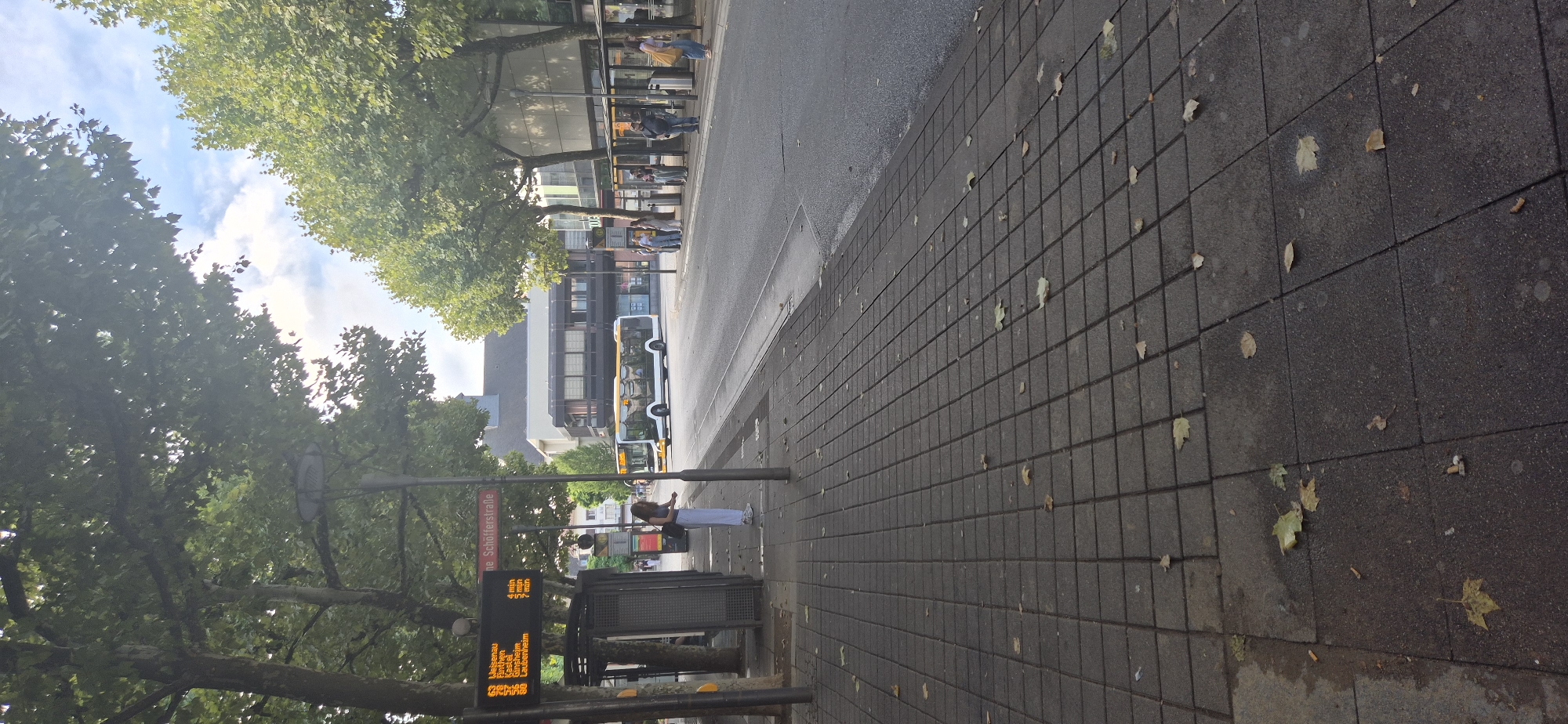 The image depicts a street scene in Mainz, Germany, specifically at the "Schöfferstraße" bus stop. In the foreground, a textured, paved sidewalk with scattered autumn leaves and cigarette butts is visible. A bus shelter with a digital display board showing bus routes and arrival times stands on the left. The display board indicates route 63 to "Weisenau" arriving in 4 minutes, and route 63 to "Laubenheim" arriving in 7 minutes. A woman in white pants and a dark top is standing near the bus shelter, looking at her phone.
Further down the street, a yellow and white bus is driving past. Several people are waiting at a bus stop on the right side of the street. Buildings with awnings and trees line the street, with a cloudy sky overhead. The overall impression is of a typical urban public transportation hub on a slightly overcast day.