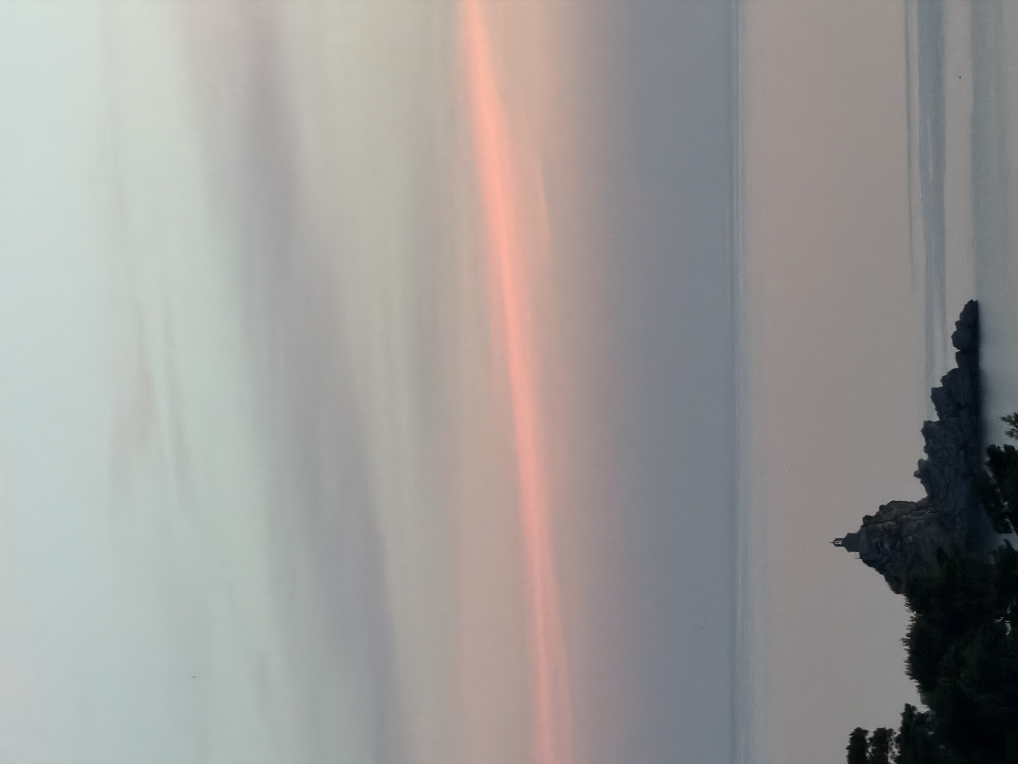 The image depicts a serene seascape at dusk or dawn. The sky is a gradient of soft blues and pinks, with a delicate horizontal band of pink cloud stretching across the mid-horizon. Below the sky, the calm sea reflects the muted colors of the atmosphere.
In the foreground, a cluster of dark green trees frames the lower left portion of the scene. To the right of the trees, a rocky outcrop rises from the water. Perched atop this outcrop is a small, dark structure, possibly a church or monument, with a distinct bell tower. The rocky island appears to be part of the coastal landscape of Petrovac na Moru, Montenegro, given its characteristic Mediterranean appearance with rugged coastline and proximity to the sea. The water is smooth and still, with a faint reflection of the sky. In the distance, a small, dark speck on the water might be a boat or a seabird. The overall impression is one of tranquility and natural beauty.