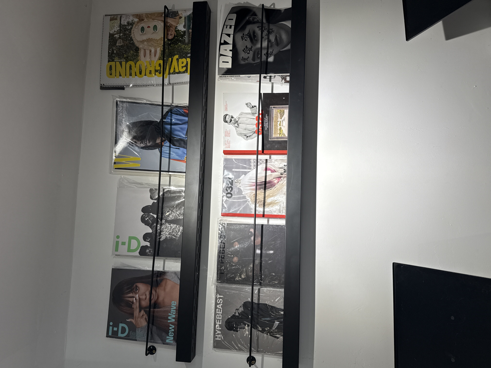 The image shows two black magazine racks mounted on a white wall. The rack on the left contains magazines with "i-D" and "Playground" displayed. One magazine features the title "New Wave." The rack on the right displays magazines with "DAZED" and "HYPEBEAST" titles. Some magazines are covered in plastic sleeves. The overall setting appears to be an interior space with neutral lighting. The presence of magazines suggests a location that may be related to fashion, design, or lifestyle.
Given the types of magazines displayed (i-D, Dazed, Hypebeast), it could be in a trendy shop, a design studio, or even someone's personal collection. Since Jing'an District is known for its fashionable spots, it aligns well with the content.
No people are visible in the image.