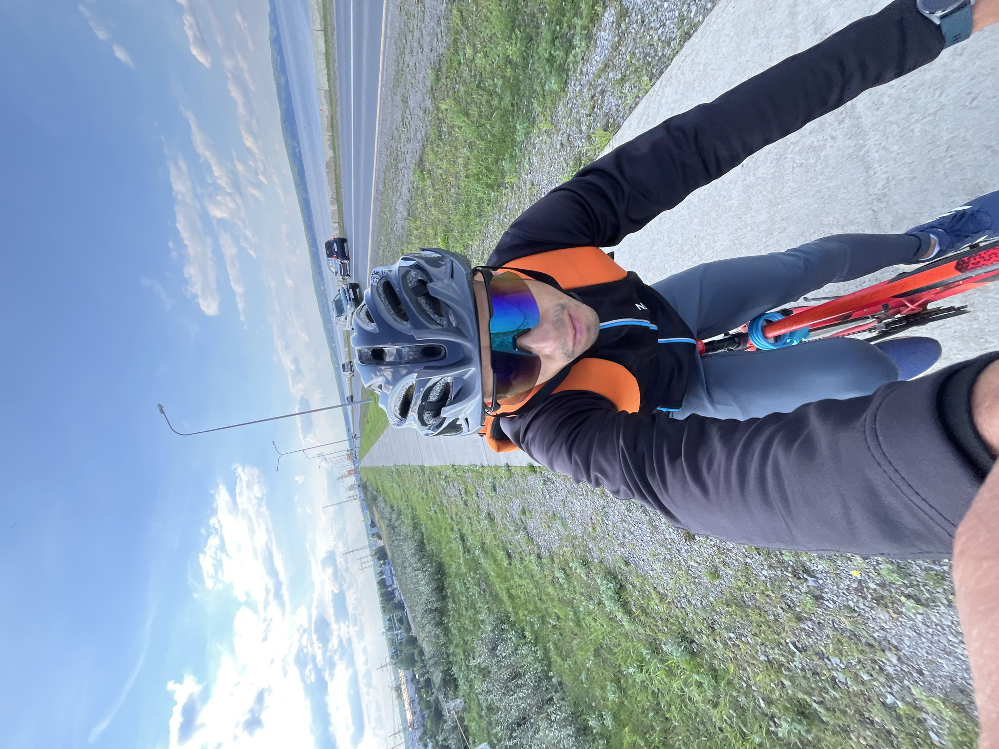 A person wearing a black jacket, grey pants, a grey helmet, and multi-colored sunglasses is riding a red bicycle on a paved path next to a road. The person is wearing an orange backpack. The road has cars driving on it and is bordered by a body of water and a hill in the distance. The sky is blue with scattered clouds. Power lines and industrial buildings are visible on the left side of the image. The photograph appears to be taken from the perspective of someone riding the bicycle, possibly a selfie. The location is the embankment of the Kama River in the city of Chaikovsky, Russia.