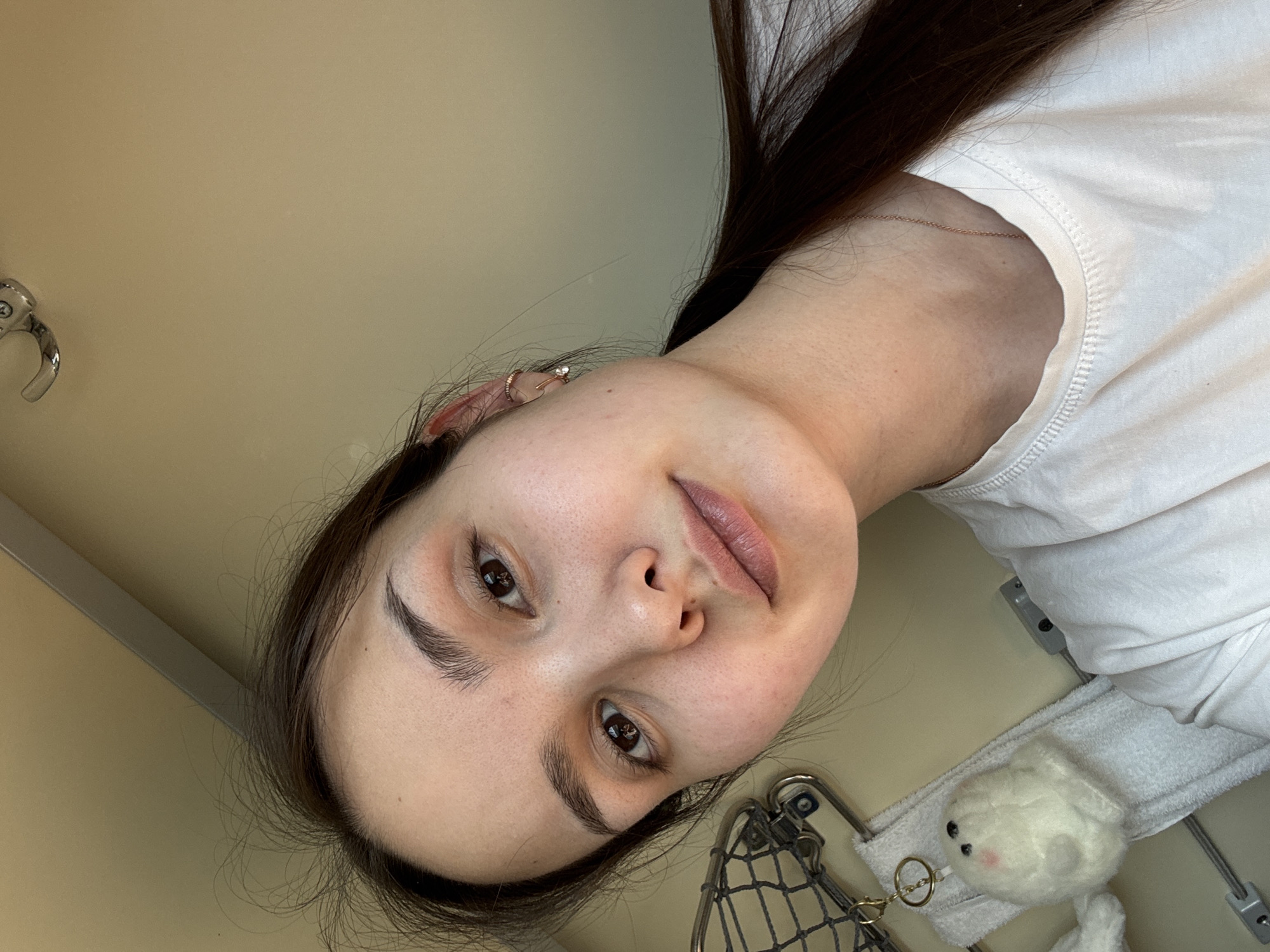 A young adult female is depicted in a close-up selfie, situated within what appears to be a train compartment, likely in or passing through the city of Kalachinsk, Russia. The subject has a light complexion, dark brown hair pulled back, and dark brown eyes, looking directly at the camera with a neutral expression. She wears a white crew-neck t-shirt. Visible accessories include a clear-stoned stud earring in her right earlobe and a small gold-toned hoop in her right upper ear cartilage. A thin, gold-toned chain necklace is visible around her neck.
The background consists of light beige wall panels. To the left, a metallic, grid-patterned overhead luggage rack is partially visible. Draped below the rack is a white terry cloth towel. Attached near the towel is a small, white, spherical plush keychain resembling a seal, featuring small black eyes and faint pink cheeks. A small, chrome-colored hook is affixed to the ceiling in the upper portion of the frame. The lighting is even, suggesting ambient indoor illumination.
