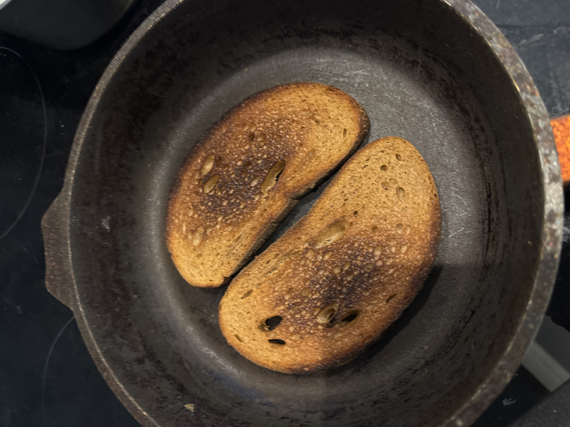 The image presents a top-down view of two slices of toasted bread resting inside a dark, well-seasoned cast iron skillet. The bread appears to be a rustic variety, possibly sourdough, characterized by an irregular shape and an open crumb with visible holes and some darker specks, which might be seeds. Both slices exhibit significant browning, with several areas showing a darker, charred appearance, indicating they are heavily toasted or slightly burnt.
The cast iron skillet is dark and has a textured, matte finish, suggesting it is well-used. It sits on a black stovetop, likely an electric or induction surface, with faint circular outlines of burners visible beneath and to the left of the pan. A small portion of an orange or reddish object, possibly a pot holder or handle, is visible near the upper right edge of the pan. The scene depicts a simple cooking activity in a kitchen environment, focused on the preparation of toasted bread. There is no visible text or identifiable people, and the time of day and weather are not discernible.