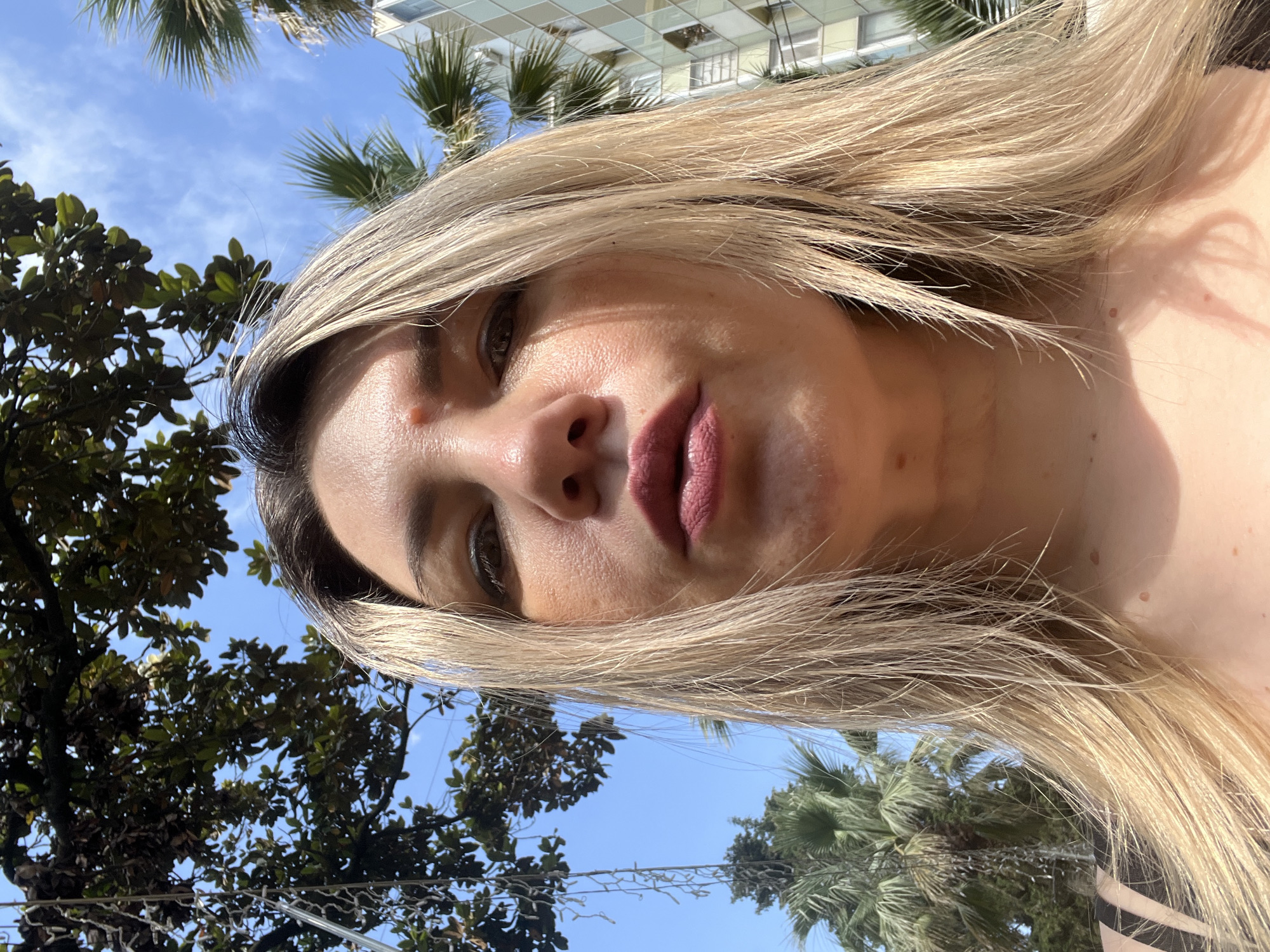 A close-up, low-angle selfie of a blonde woman with light skin, wearing dark lipstick. She is outdoors, with a blue sky and leafy trees visible. The background includes palm trees and a modern building. The sunlight casts shadows on her face. The location is Sochi, Russia.