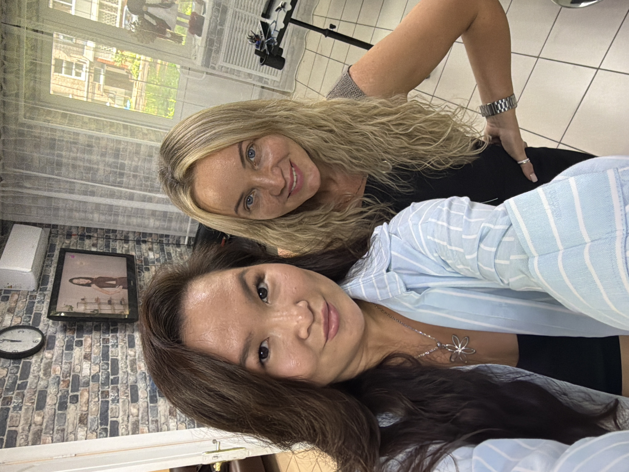 Two women are taking a selfie in a room that appears to be a hair salon. The woman on the left has long, dark brown hair and is wearing a light blue and white striped button-down shirt over a black top and a silver flower pendant necklace. The woman on the right has long, wavy blonde hair and is wearing a black top and a silver watch on her left wrist. She is smiling and looking at the camera. In the background, there is a brick wall with a clock and a television screen displaying a woman. There are also windows with sheer curtains and a trolley with hair styling equipment. The floor is tiled. The overall setting suggests an indoor commercial establishment. The image is taken from a slightly high angle.
