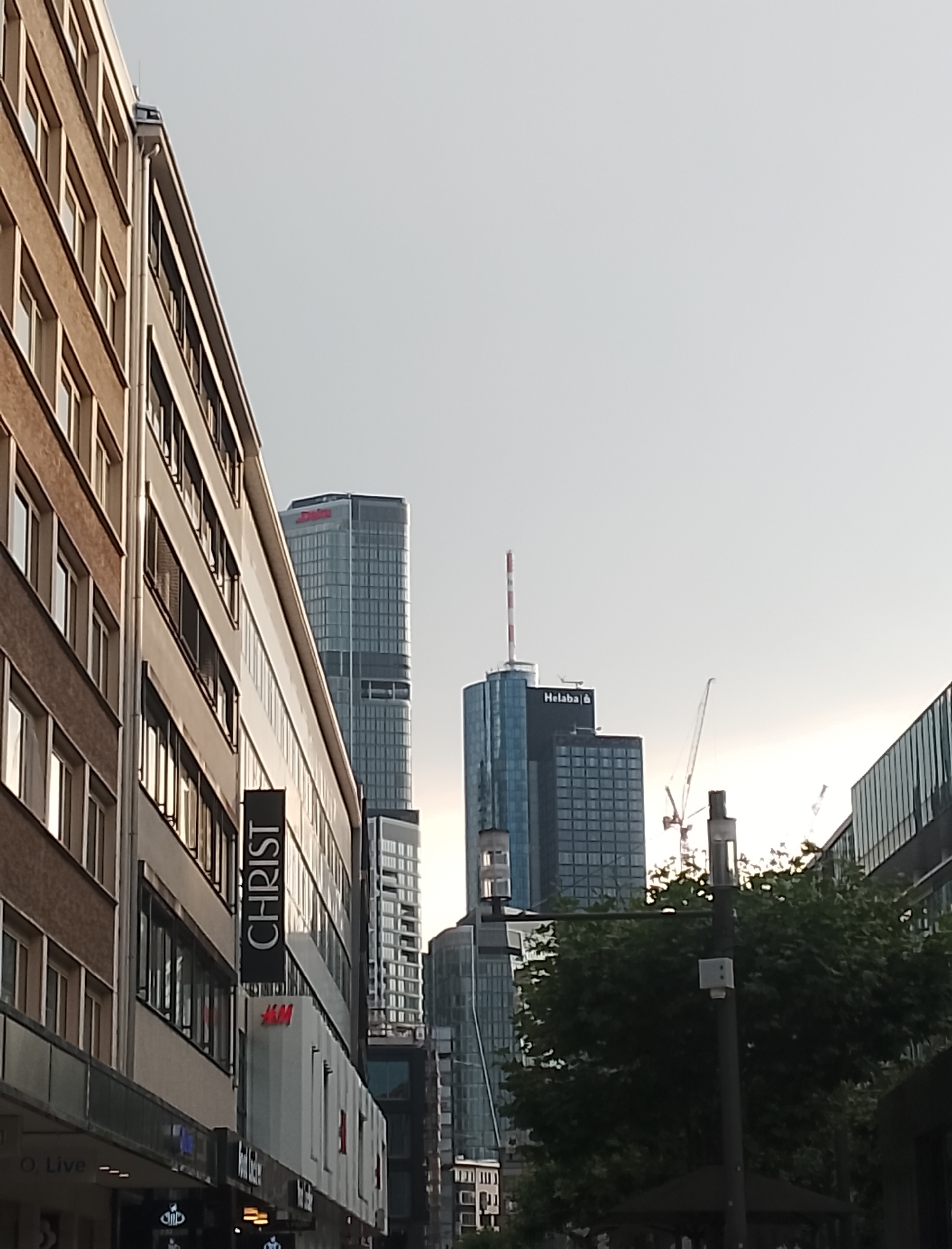 An outdoor, eye-level shot captures a city street scene. On the left, a brick building's facade, featuring multiple rows of windows, extends upwards. To its right, a modern building with a glass facade and horizontal window bands is visible. Further in the background, a tall, slender skyscraper with a distinctive red and white antenna at its apex dominates the skyline. Another skyscraper, identified by the "Helaba" logo, stands to the right of the first. The sky is clear and bright, suggesting daytime. Several construction cranes are also present in the background, indicating ongoing development. In the foreground, a dark banner with the white text "CHRIST" hangs vertically. Below it, signage for "H&M" is visible. Streetlights and trees are also present in the scene.