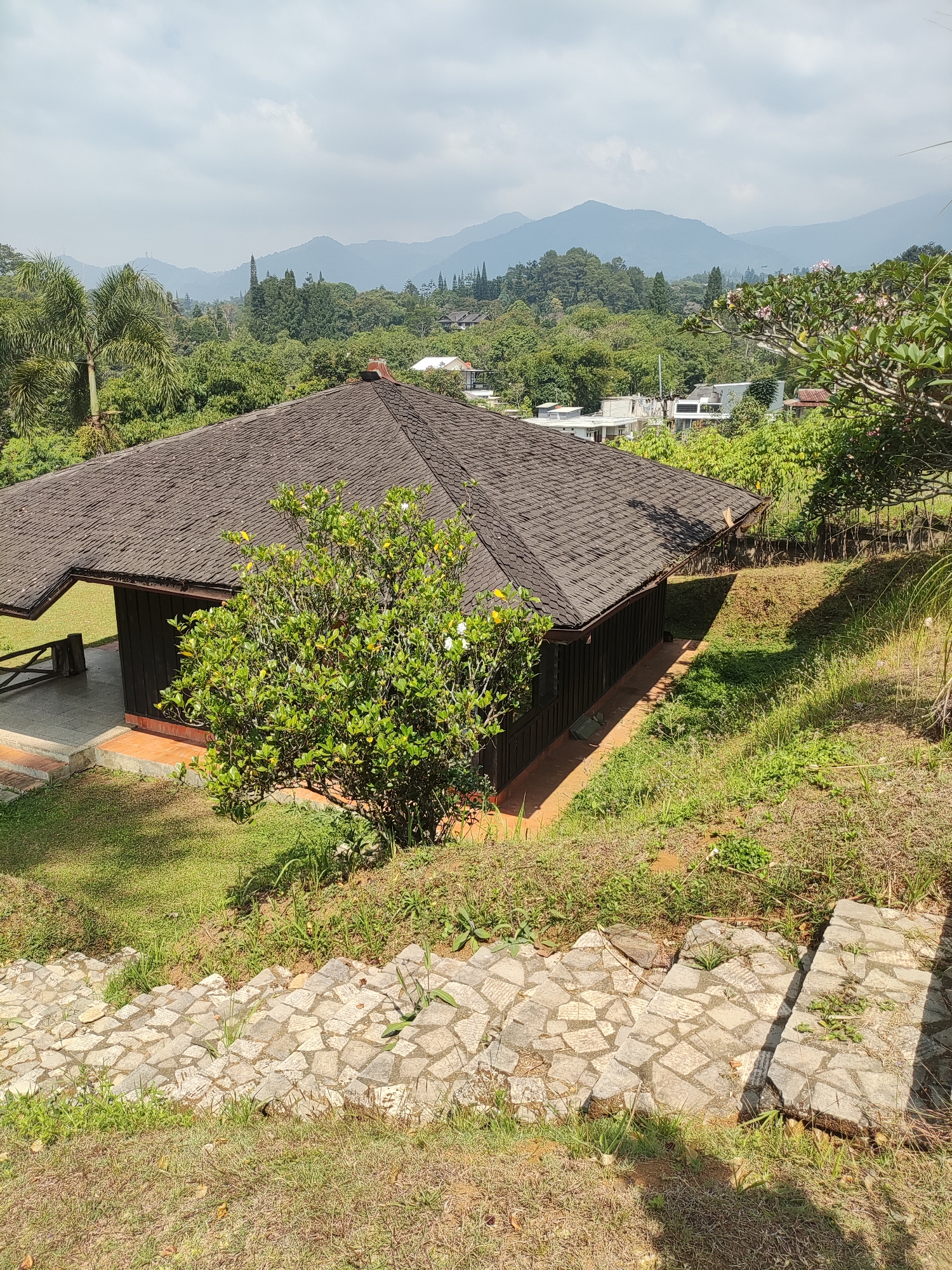 A high-angle outdoor shot depicts a bungalow with a dark brown, shingled roof situated on a grassy slope. The bungalow features dark wooden walls and an open veranda with a concrete floor. A lush green tree with white flowers stands in front of the bungalow. In the foreground, a stone staircase descends the slope. Beyond the bungalow, a dense forest covers the landscape, leading to distant, hazy mountains under a cloudy sky. The location is identified as Cisarua, Indonesia.