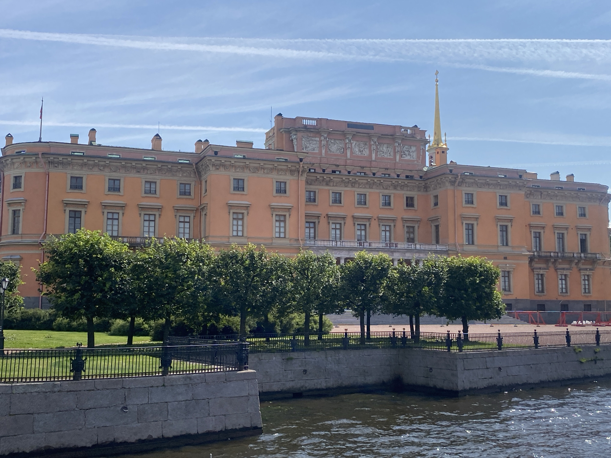 The image shows a waterfront view of the Mikhailovsky Castle in Saint Petersburg, Russia. The castle is a large, orange building with multiple windows and decorative elements on its facade. It is topped with a prominent golden spire. In front of the castle is a well-maintained lawn with several rows of neatly trimmed trees. A black metal fence separates the green space from the canal, which occupies the foreground of the image. The water in the canal is dark and rippled. The sky is blue with thin, wispy white clouds. The scene is calm and picturesque, showcasing a notable architectural landmark of the city.