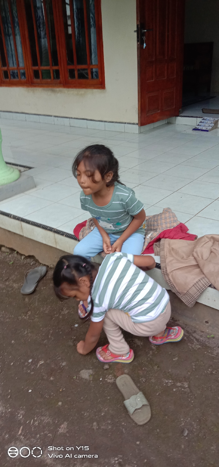 The scene depicts two young children situated outside a residential building in Ranugedang, Indonesia, on a patio adjacent to a dirt ground. The building features a light-colored wall, a large wooden window frame with dark curtains visible through its panes, and a partially open wooden door with a key hanging from the handle. A light green pillar is visible on the left side of the tiled patio.
An older child, appearing to be a girl, is seated on the tiled patio, resting on a red and plaid patterned cushion. She wears a light green striped short-sleeve shirt with a white graphic and light blue pants. Her dark hair is loose, and her eyes are partially closed, with her mouth slightly open. Beside her, on the patio, is a folded brown garment.
A younger child, appearing to be a girl, is crouched on the uneven, damp dirt ground immediately in front of the patio. She wears a white and green striped short-sleeve shirt, light brown pants, and multi-colored flip-flops. Her dark hair is pulled back with pink ties. She is looking downwards towards the ground, near her hands.
On the dirt ground, a single grey flip-flop is positioned to the left of the younger child, and a single light green slide sandal is to her right. The overall environment suggests an outdoor residential area.