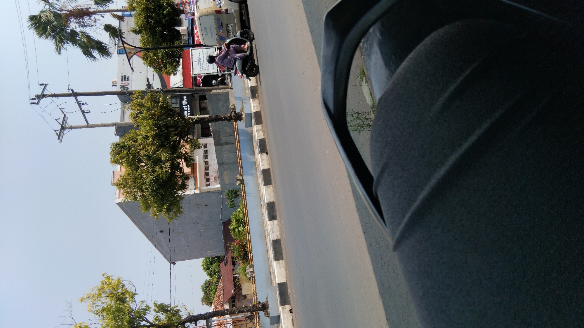 A person wearing a purple headscarf and tunic rides a dark-colored scooter with a white rear fender along a paved road in Batang, Indonesia. The scooter is moving away from the viewer, slightly to the right. In the background, there is a light-colored commercial building with signage that reads "House of Diva BEAUTY SALON". A white van with the word "TASKO" visible on its side is parked next to the building. Overhead, utility poles with wires stretch across the clear blue sky. Trees with green foliage line the street, and a concrete barrier with black and white stripes separates the road from a sidewalk and a grassy area. The foreground of the image is dominated by the black textured surface of a vehicle's dashboard or seat, with a curved line suggesting the edge of a mirror or windshield. The overall scene depicts a typical urban streetscape on a sunny day.