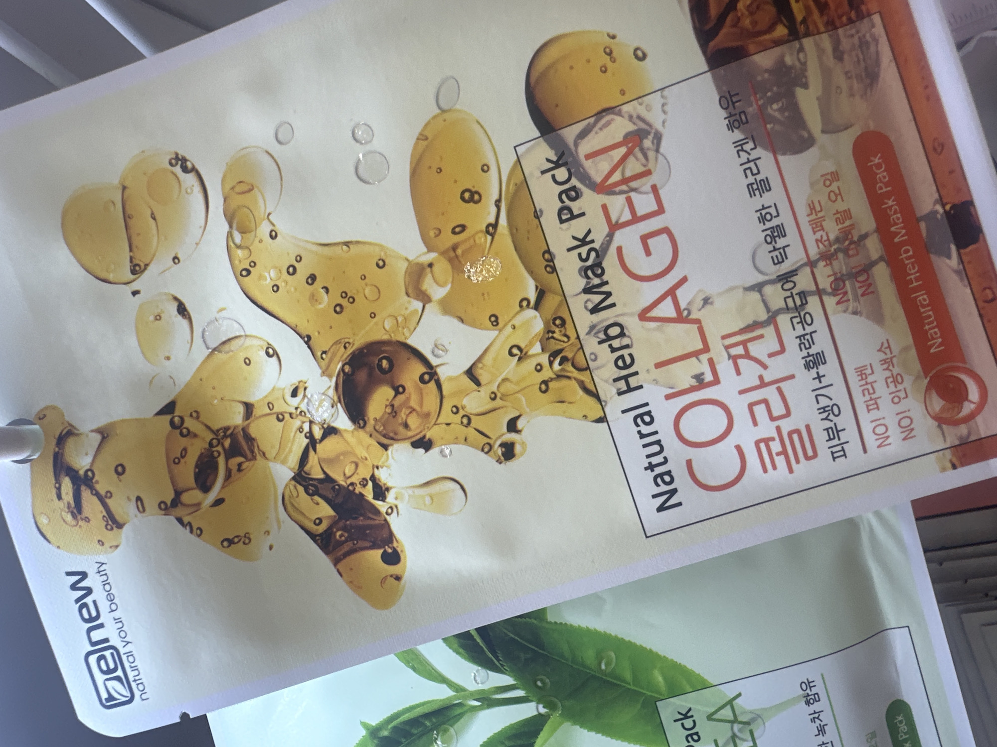 The image displays a close-up, slightly angled view of two skincare product packages. The primary focus is on a package for "be new natural your beauty" facial mask, which features a prominent graphic of golden, viscous liquid with bubbles and streaks. The text "Natural Herb Mask Pack COLLAGEN" is clearly visible in large, red lettering. Below this, there is Korean text and smaller English text. To the left of this package, a portion of another package is visible, showing green tea leaves and droplets. The background is neutral and appears to be a retail setting. The image was taken in Bac Nha Trang Ward, Vietnam.