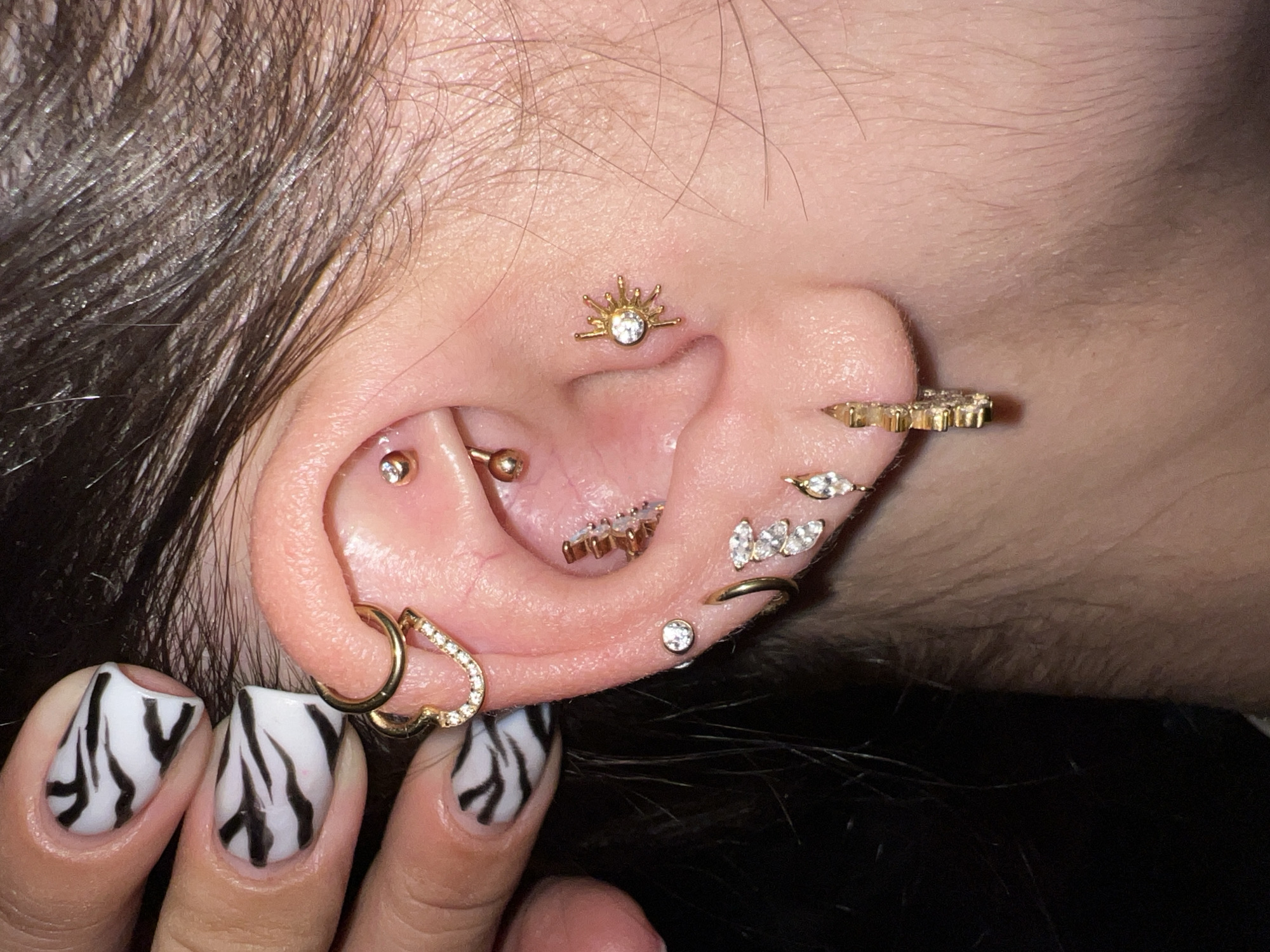 A close-up shot features a woman's ear adorned with multiple gold-colored piercings, including a sunburst stud in the conch, a row of marquise-shaped gems in the helix, and a heart-shaped hoop at the lobe. Her nails are decorated with a black and white zebra-stripe pattern. The background is dark, and parts of her dark brown hair are visible. The image does not show or mention anything related to the city of Noviy, Russia.