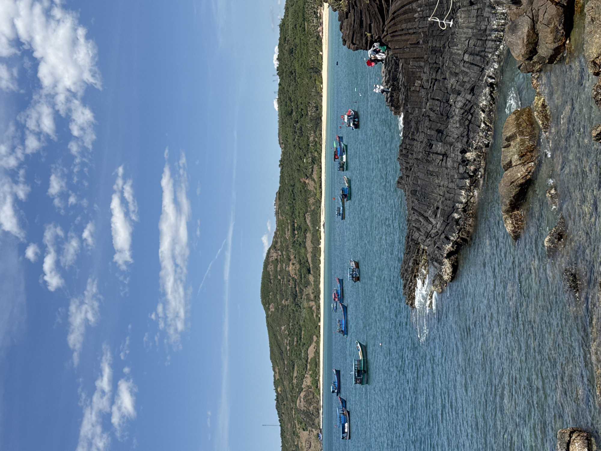 The media file displays a scenic coastal view at Phú Lương, Vietnam. The scene is dominated by a bright blue sky with scattered white clouds, suggesting fair weather during daytime. The main subjects are a row of small boats floating on clear, turquoise water. The boats are of various colors, including blue, green, and red, and appear to be fishing or leisure vessels.
In the foreground, dark, rugged volcanic rocks form a natural barrier along the coastline. Some of these rocks have a columnar jointing pattern, characteristic of cooled lava flows. A few people are visible on top of these rocks, observing the seascape or preparing for activities. The water near the rocks shows some gentle waves and splashes.
Behind the boats, a narrow, sandy beach stretches along the coast, backed by a lush green, tree-covered hillside. The hillside rises steeply, creating a dramatic backdrop to the tranquil beach and water. In the distance, the land merges with the horizon under the vast sky.
The overall impression is one of a peaceful day at a tropical coastal location. The combination of the blue sky, clear water, green hills, and the moored boats creates a picturesque and inviting atmosphere. There is no visible text in the media file.