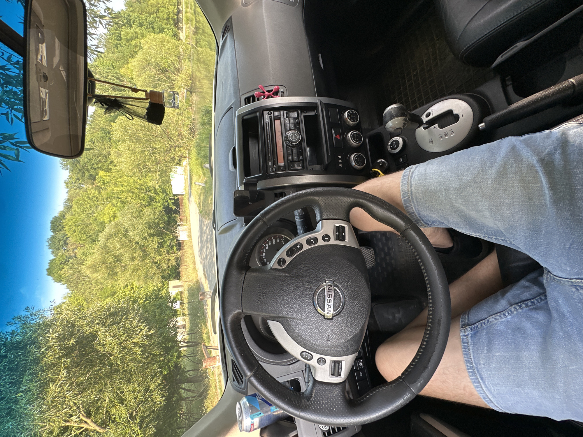 The image provides an interior view from the driver's seat of a car, looking forward. The driver, wearing denim shorts, has their legs visible. The steering wheel, dashboard, and center console of a Nissan vehicle are prominent. On the dashboard, a car stereo and air conditioning controls are visible, along with a car air freshener. A can of soda is placed in a cupholder. Outside the car, through the windshield, a natural landscape is seen, with trees lining a body of water, possibly a lake or river, under a clear blue sky. The location is identified as Makiv, Ukraine, though no specific landmarks within the city are directly visible. The scene appears to be a snapshot of a moment while parked or driving through a scenic, possibly rural, area near Makiv.