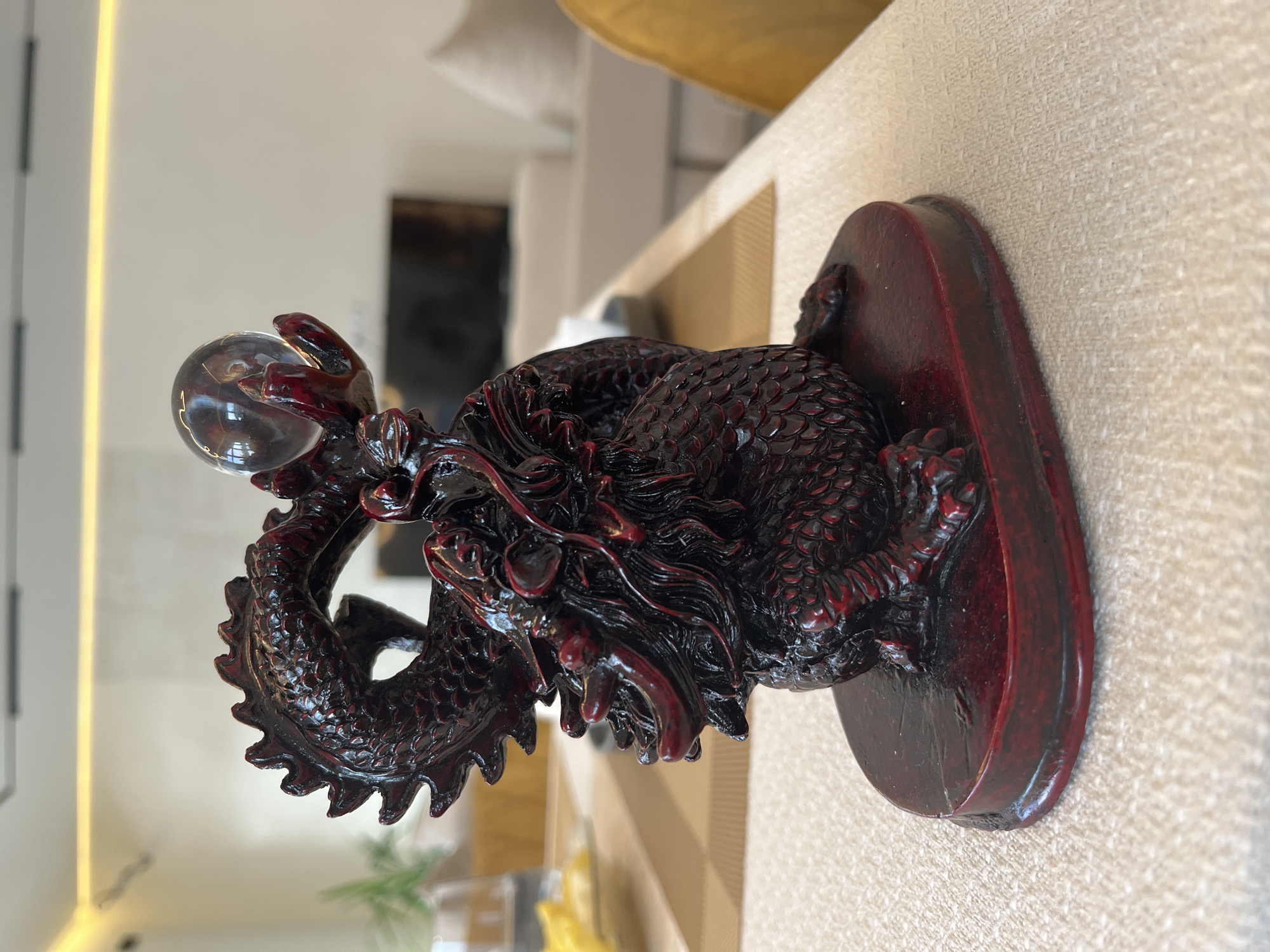 An intricately carved dark reddish-brown dragon figurine, appearing to be resin or lacquered wood, is depicted holding a clear spherical crystal orb in its raised claws. The dragon, detailed with scales and a textured mane, rests on a matching oval-shaped base. This figurine is situated on a light beige woven tablecloth, with a rectangular brown placemat visible beneath it and extending to the left. In the blurred background to the left, a clear glass and yellow objects on a white dish are partially discernible. The wider background reveals a modern indoor setting with light-colored walls, a linear warm LED light strip along the ceiling, and out-of-focus light-colored furniture (possibly a sofa) and a dark screen. A yellow chair back is visible on the far right. The scene is located in Новая Чернушка, Russia.