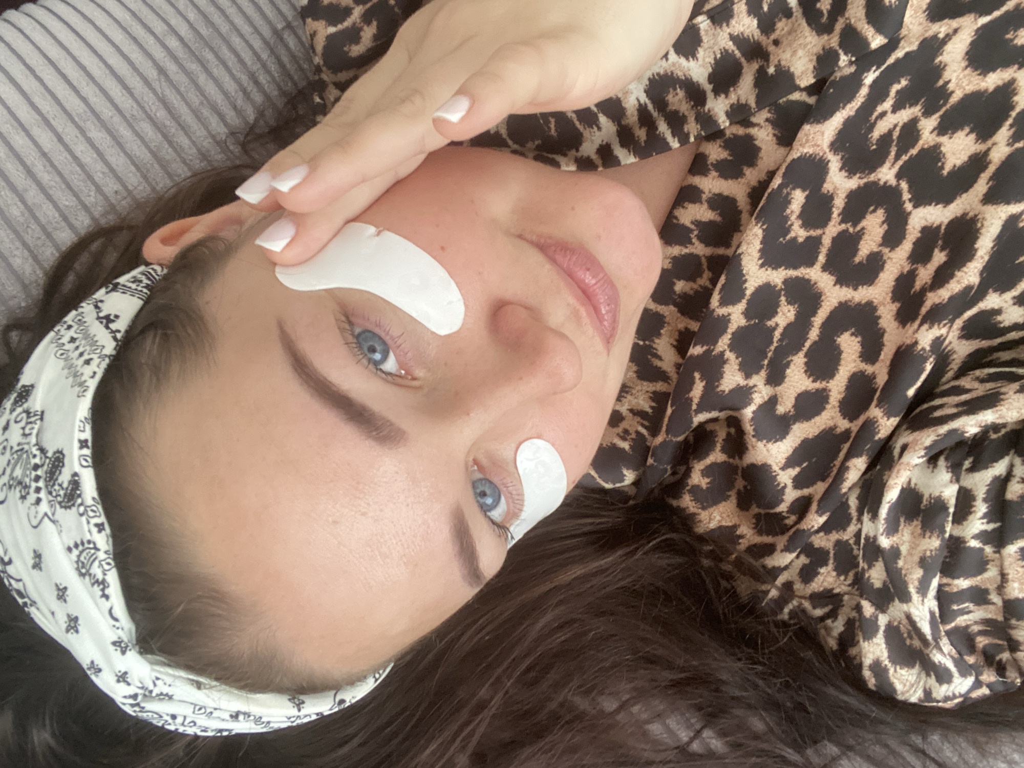 A young woman is captured in a close-up, high-angle shot, lying down and looking upwards towards the camera. She has blue eyes, defined eyebrows, and appears to be undergoing a beauty treatment, as indicated by the white, crescent-shaped under-eye patches positioned beneath each eye. Her lips are adorned with a light pink gloss, and her skin shows a few subtle freckles.
Her dark brown hair is held back from her face by a white bandana featuring a black paisley pattern. She is wearing a top or pajamas with a distinct brown and black leopard print. One hand, displaying neatly manicured nails with white tips, rests gently near her forehead. The background consists of a striped grey and white textile, likely a pillow or blanket, partially obscured by the leopard print fabric of her clothing. The overall scene suggests a relaxed atmosphere, possibly at home or in a salon, located in Sochi, Russia. No visible text is present in the image.