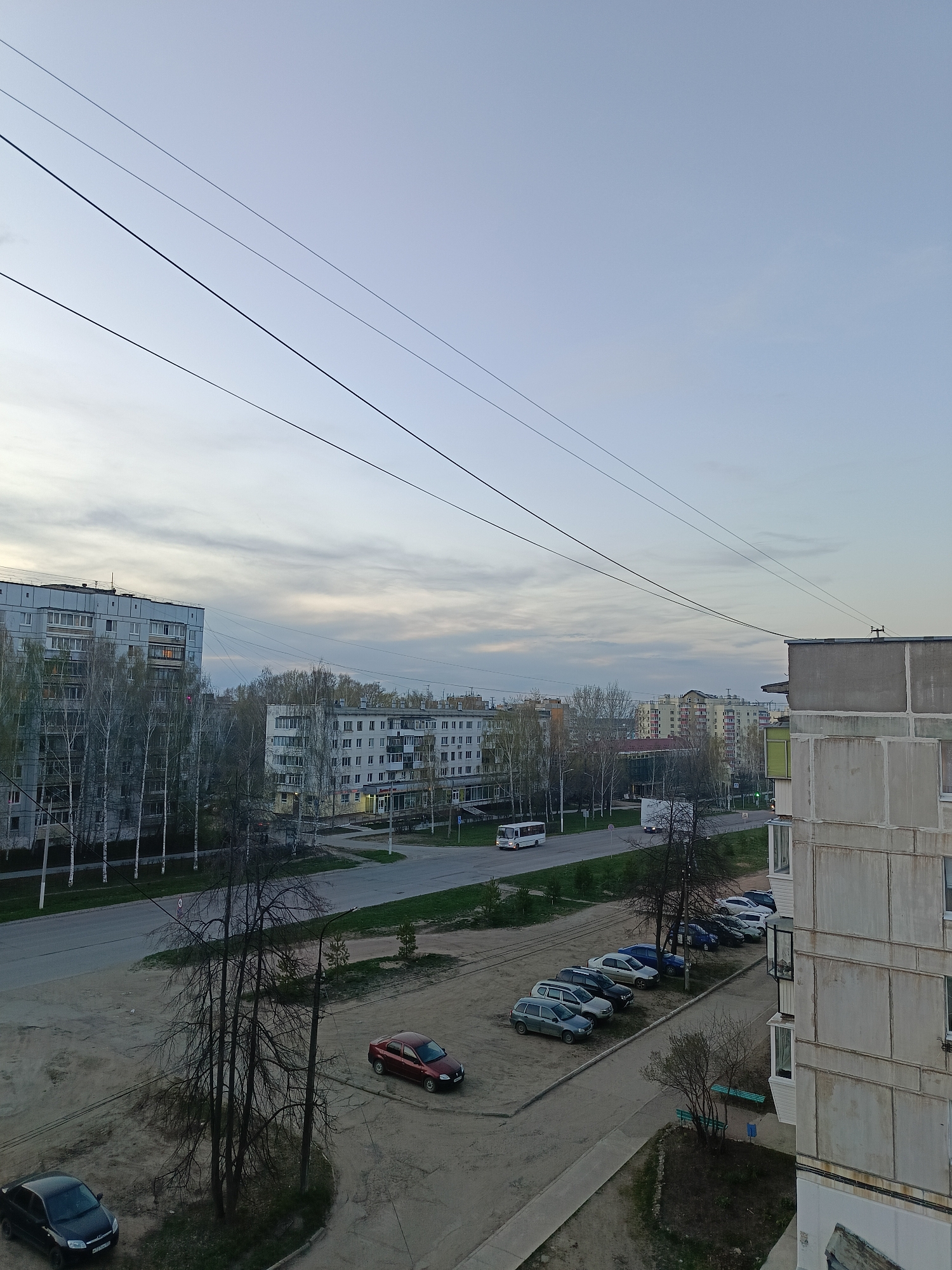 An elevated view depicts a typical residential area in Chaikovsky, Russia, under a sky obscured by dull, overcast lighting. In the foreground, a paved parking lot is adjacent to a weathered, Soviet-era apartment building, displaying stark, concrete paneling. The parking area contains various parked cars of different models and colors. Beyond the parking area, a street stretches across the midground, featuring sparse greenery and pedestrian paths, punctuated by small trees. A white bus and another vehicle travel on the street. In the background are more Soviet-era apartment buildings. Telephone wires stretch across the upper portion of the frame. No people are clearly visible.