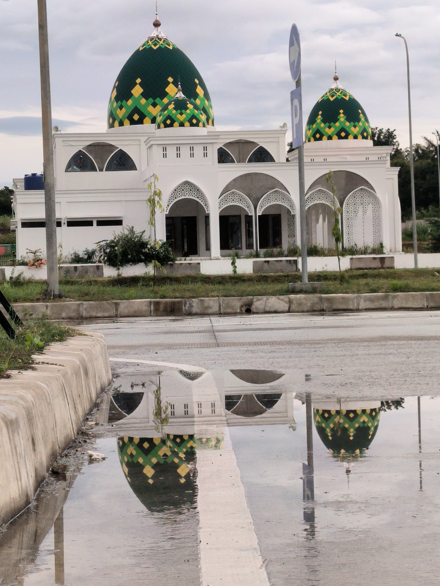 A white mosque with striking green and yellow domes is the central subject of this vertical photograph, prominently featured and reflected in a roadside puddle. The mosque's architecture includes a large main dome and a smaller secondary dome, both intricately decorated with a geometric pattern of green, yellow, and black diamond shapes. The main building is white, featuring multiple arched windows and entrances adorned with decorative lattice work, as well as some simpler rectangular windows on its upper sections.
In the foreground, a large puddle of water on the asphalt road serves as a mirror, offering a clear reflection of the mosque's domes, part of its white facade, and a nearby utility pole. A white road marking is faintly visible within the puddle. A concrete curb separates the road from a narrow strip of grass on the left. Two grey utility poles are present; one on the far left and another closer to the mosque. A blue circular road sign, displaying a white arrow pointing right and a subtle 'P' below it, stands to the right of the mosque. The sky is overcast and uniformly grey, suggesting a cloudy daytime. The substantial puddle indicates recent rainfall, contributing to a quiet and still atmosphere. No people or vehicles are visible in the scene.