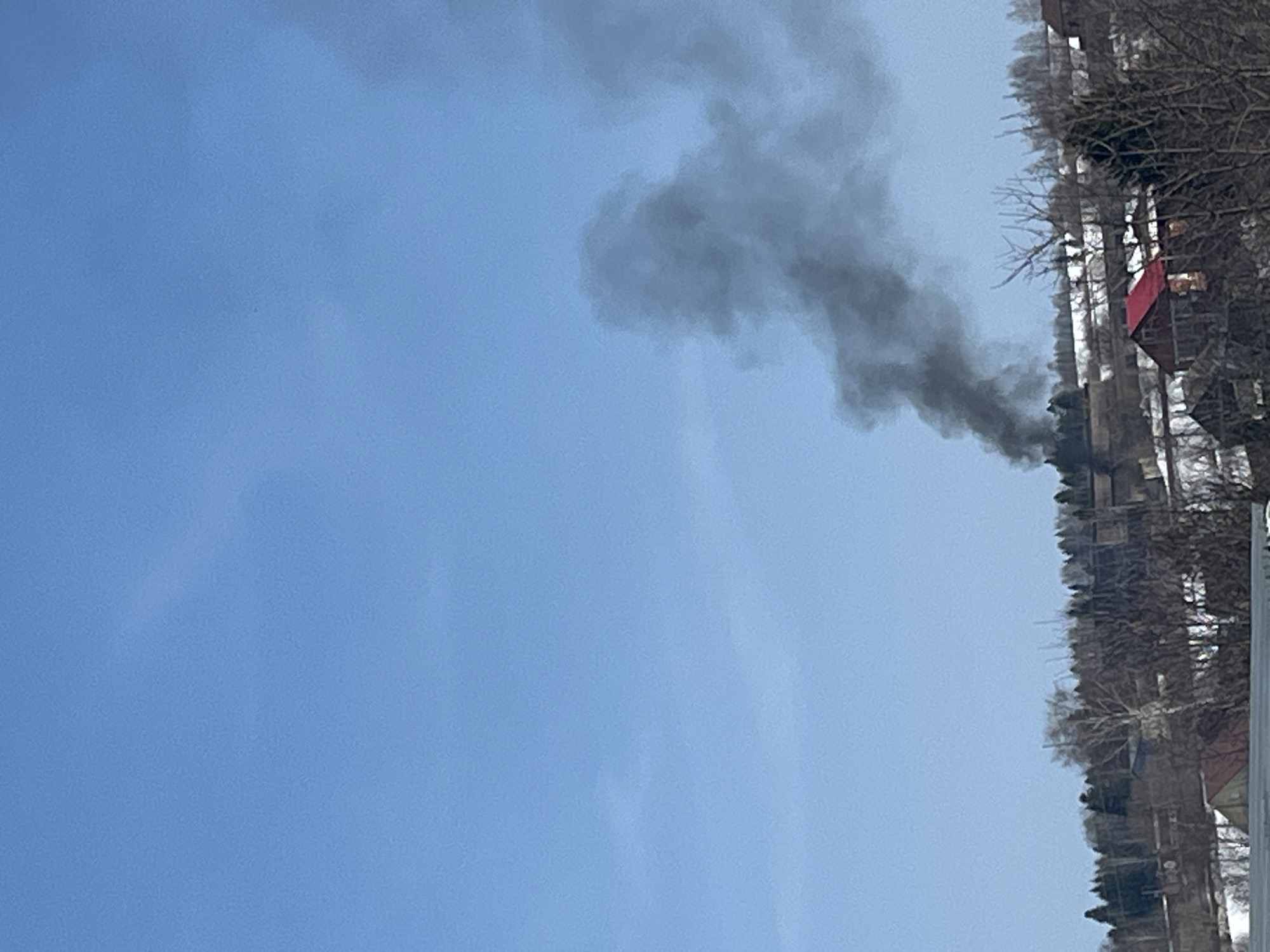 The image shows a plume of black smoke rising into the sky near Pavlovka, Russia. The buildings are visible on the right side of the image, suggesting a dense urban environment. There are trees around the area. The sky is blue, and the weather conditions seem to be relatively clear, allowing the smoke to be seen from a distance.