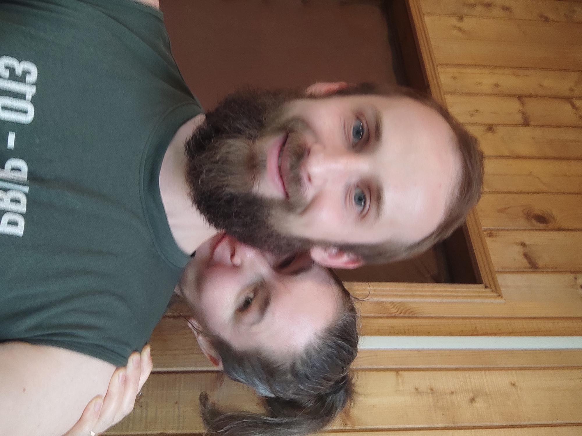 A man with a beard and a woman are standing close together indoors. The man is wearing a dark green t-shirt with some white text on it, while the woman has her hair pulled back. They both appear to be looking at the camera and smiling slightly. The background is a wooden wall. The exact location in Kratovo, Russia, cannot be determined from the visual information.
