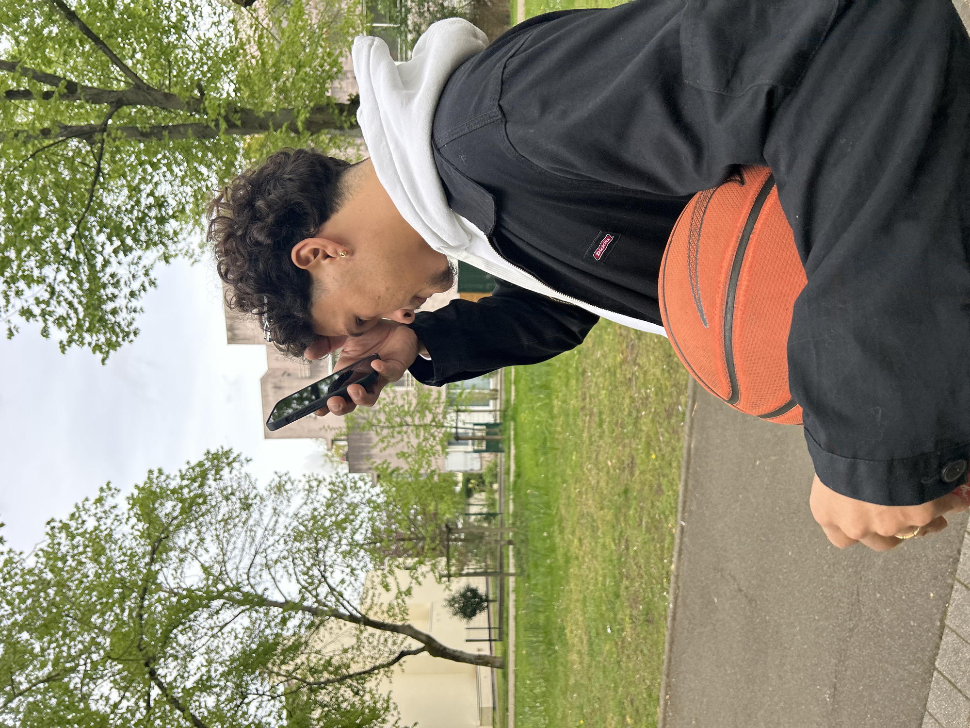 A person stands outdoors on a sidewalk in what appears to be a public park or green space in Noisiel, France. The person is holding a smartphone in their left hand and looking at the screen with their head tilted downwards. They are wearing a black jacket over a white hoodie, and an orange basketball is positioned between their left arm and body. The background includes green grass, trees with budding leaves, and buildings, suggesting an urban environment. The image is oriented in landscape mode. The brand "Dickies" is visible on the person's jacket.