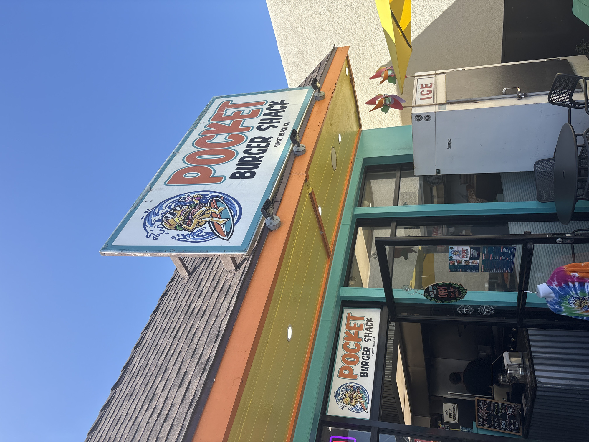The image shows the "Pocket Burger Shack" restaurant in Sunset Beach, Huntington Beach, California. The restaurant has a large sign with the name on it, plus another sign on the building window. It also has an ice cooler and a table with chairs outside.