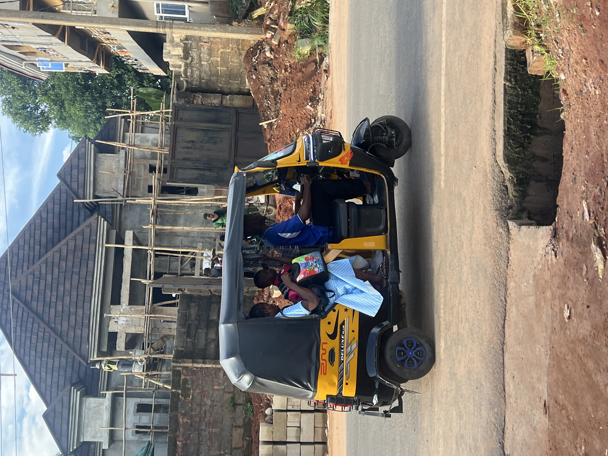 In Nkpor, Nigeria, a yellow and black auto-rickshaw is driving on a paved road. The auto-rickshaw has a black canopy and is carrying three people. In the front passenger seat, a young boy with a backpack is visible. In the rear passenger seat, a woman in a blue and white striped dress is seated. The driver, wearing a blue shirt and black pants, is operating the vehicle. The background shows a house under construction with scaffolding and roofing materials. To the right, another multi-story building with balconies is present, and a pile of reddish dirt is on the side of the road.