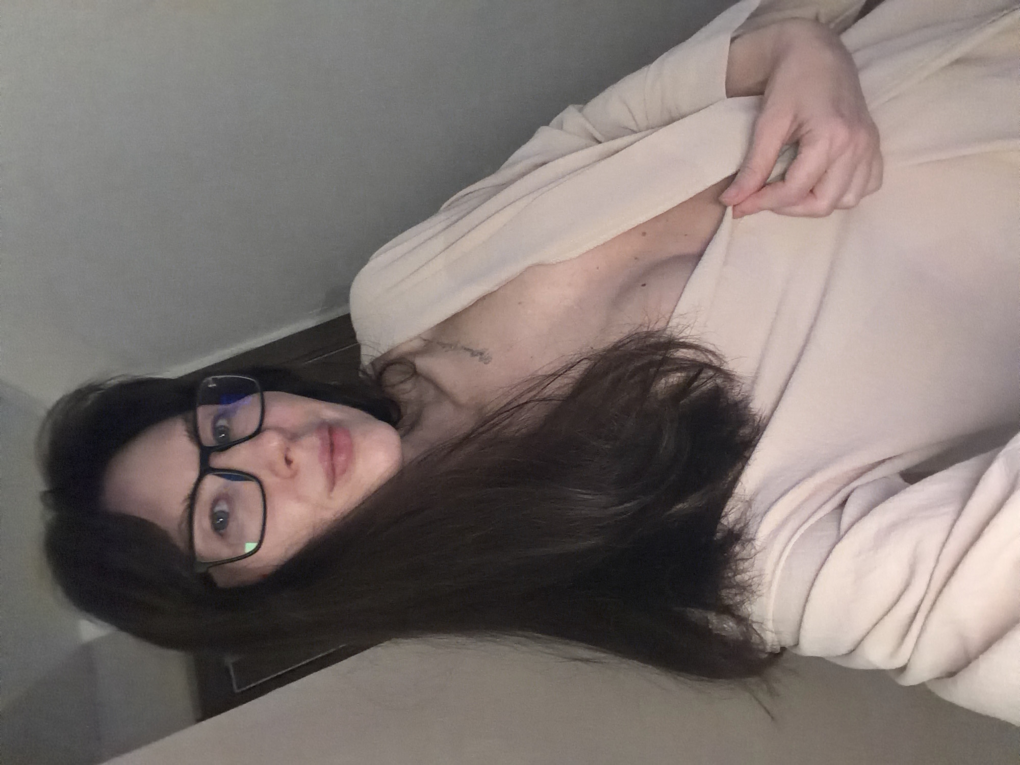 A woman with brown hair and glasses is lying down, wearing a light-colored garment. She is holding the garment slightly open near her collarbone. The location is potentially Perm, Russia, but this is not definitively confirmed by the image alone. The environment appears to be indoors, with a wall visible in the background.