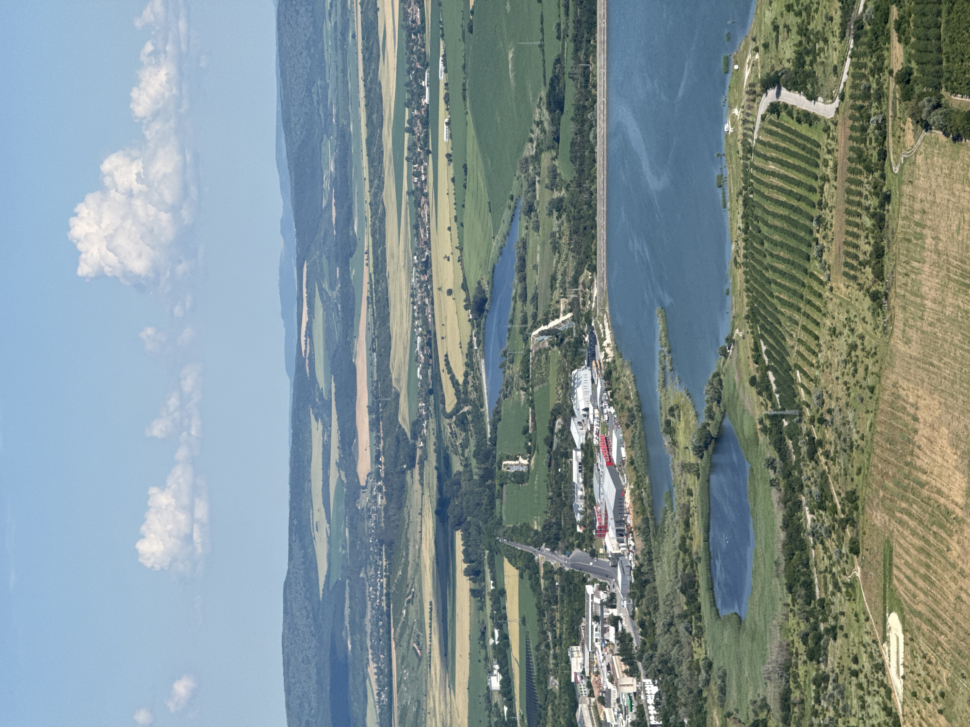 The image is an aerial view of a landscape near Shumen, Bulgaria, featuring a large body of water, agricultural fields, and some industrial or commercial buildings. In the foreground, a wide, dark blue body of water dominates the lower right portion of the image, with a smaller, irregularly shaped body of water to its left, surrounded by green reeds and vegetation.
To the left of the larger body of water, there is a settlement or industrial area with various buildings, some appearing to be warehouses or factories with red accents. A road runs through this area, connecting to the wider landscape. Beyond the buildings, a long, straight road stretches into the distance, lined with trees and leading towards a more rural area.
The middle ground of the image shows a patchwork of agricultural fields in various shades of green and yellow-brown, interspersed with lines of trees and smaller bodies of water. In the background, rolling hills covered in dark green vegetation create a layered effect, with lighter green and yellow fields visible on their slopes. The sky above is a clear blue with a few fluffy white clouds. The overall impression is a semi-rural area with significant water features and agricultural activity.